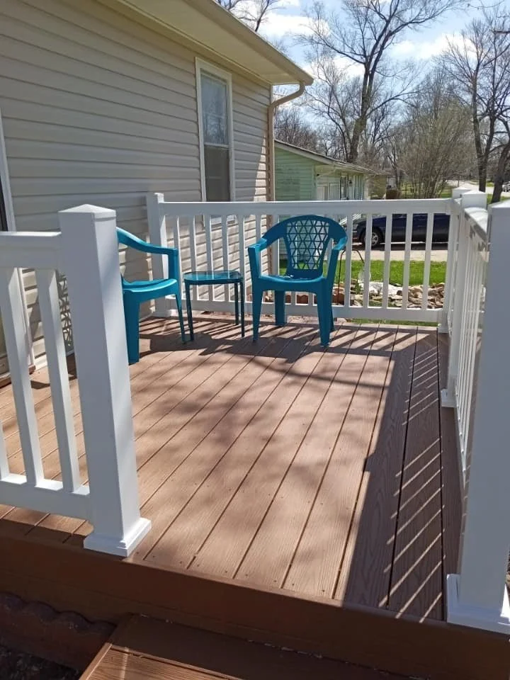 A newly built wooden porch with white railings attached to a house with beige siding. There are two blue chairs and a small blue table on the porch, with a tree and neighboring houses visible in the background.
