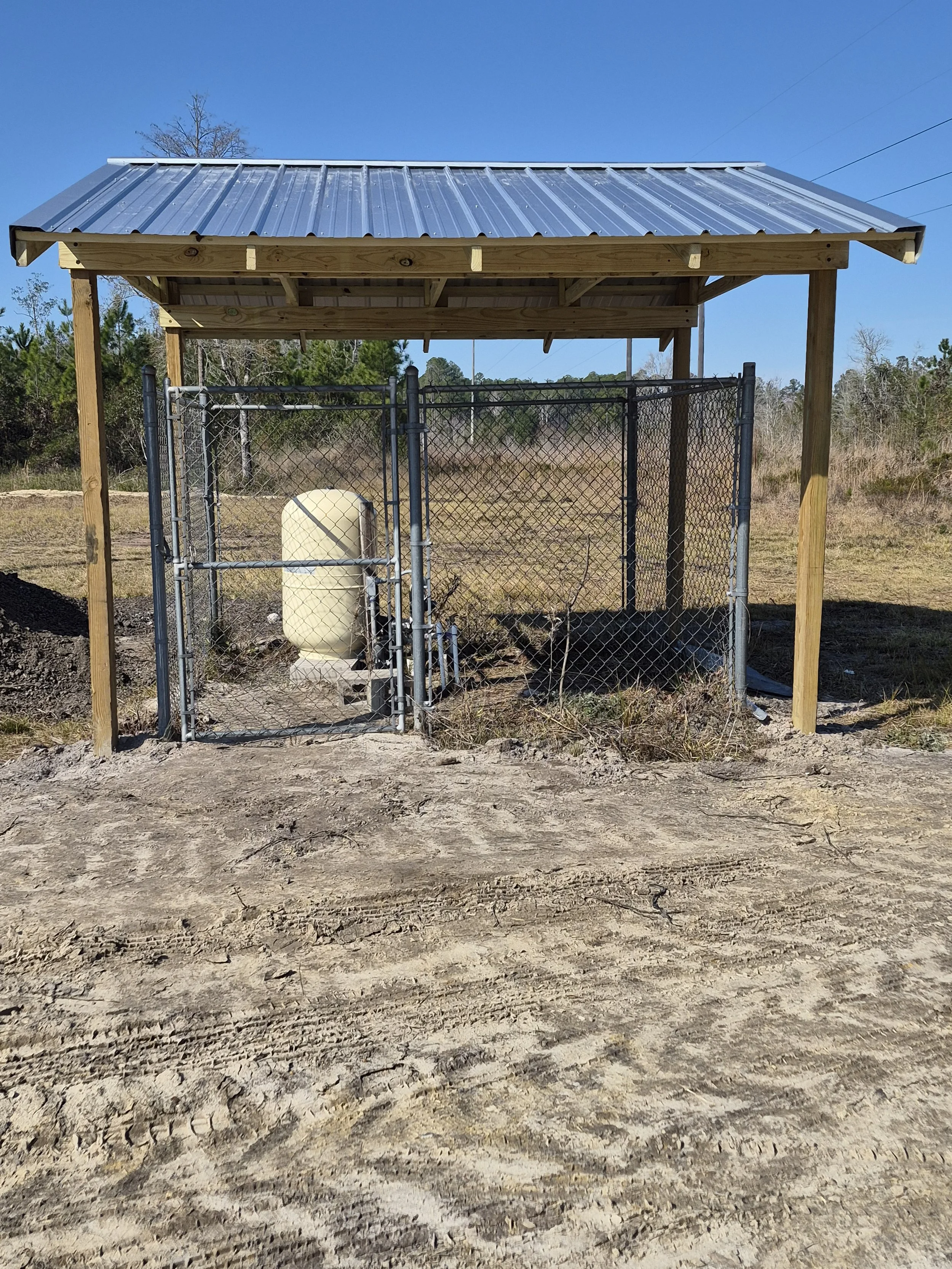 A small structure with a metal roof and chain-link fence surrounding a water tank, situated in an open, grassy area under a clear blue sky.