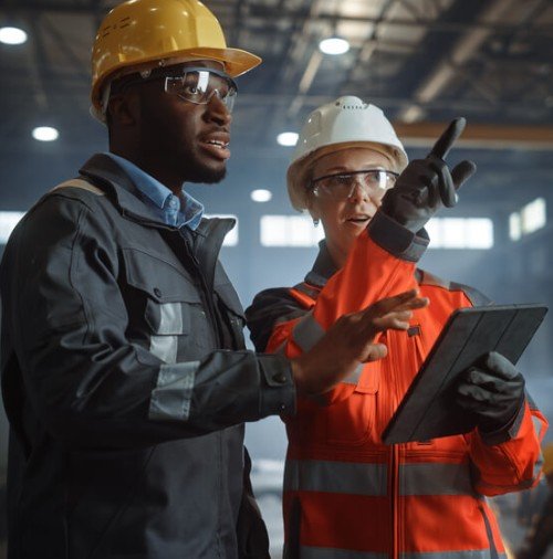 Two construction workers, a man and a woman, wearing safety helmets and protective gear, are inspecting a tablet and discussing in an industrial setting.