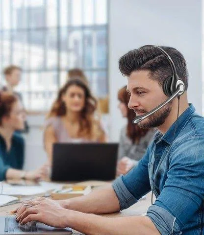 A man wearing a headset working at a computer in an office setting, with a group of blurred people in the background.