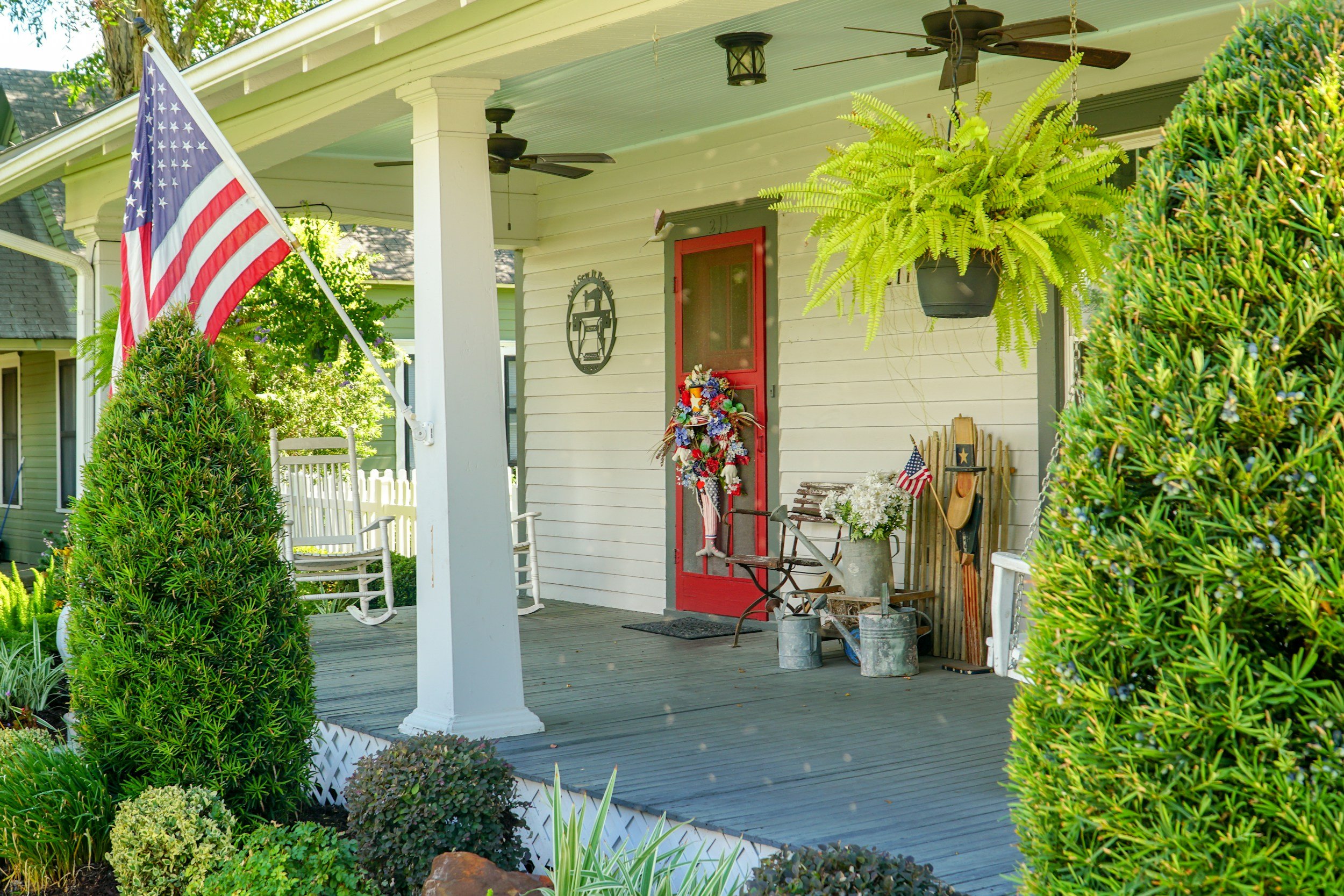 Front porch of a house decorated for the Fourth of July with an American flag, a wreath on the red door, potted plants, and patriotic decorations.