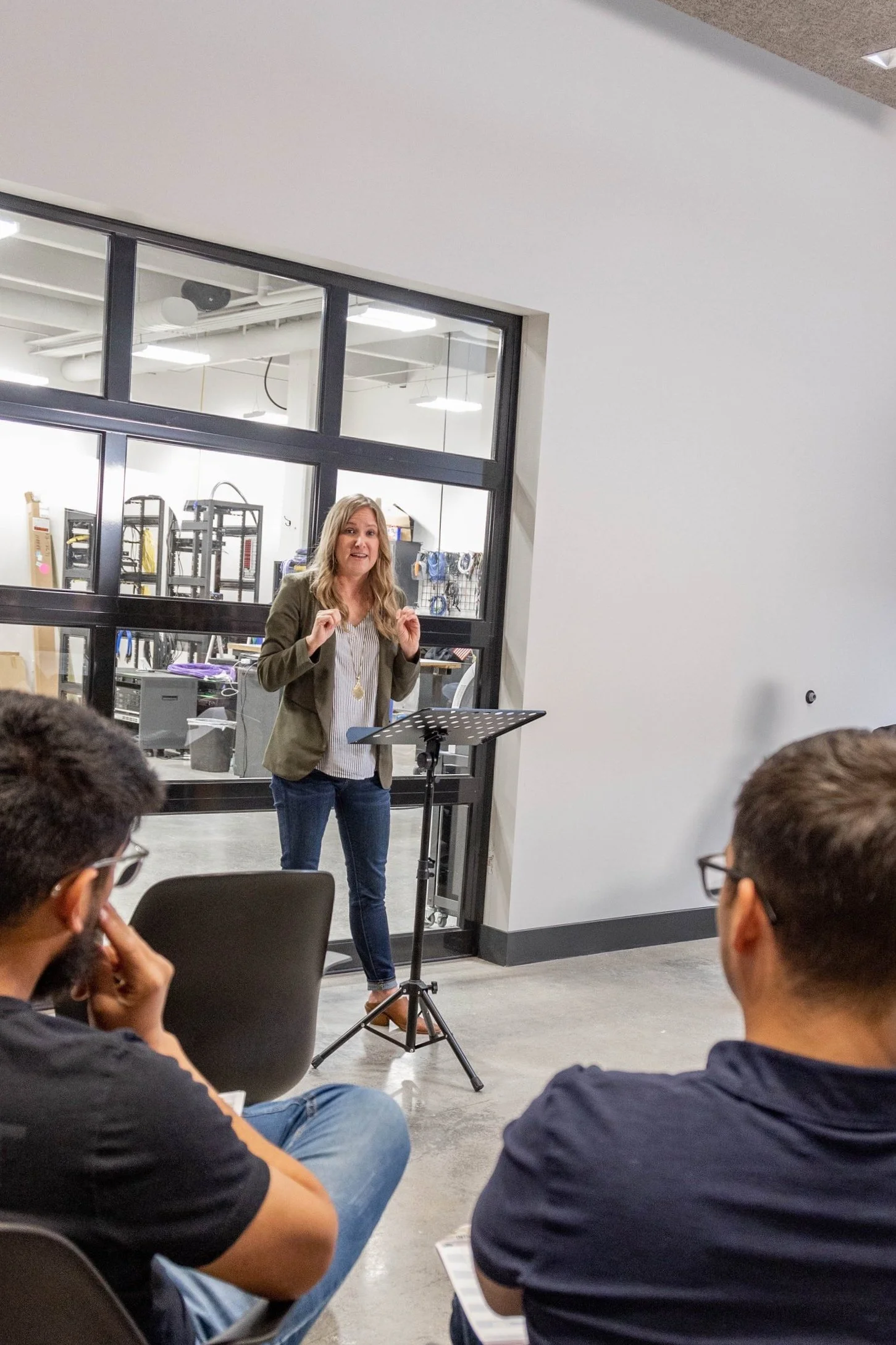 A woman giving a presentation to a small audience in a modern, industrial-style room with glass walls, and a music stand in front of her.
