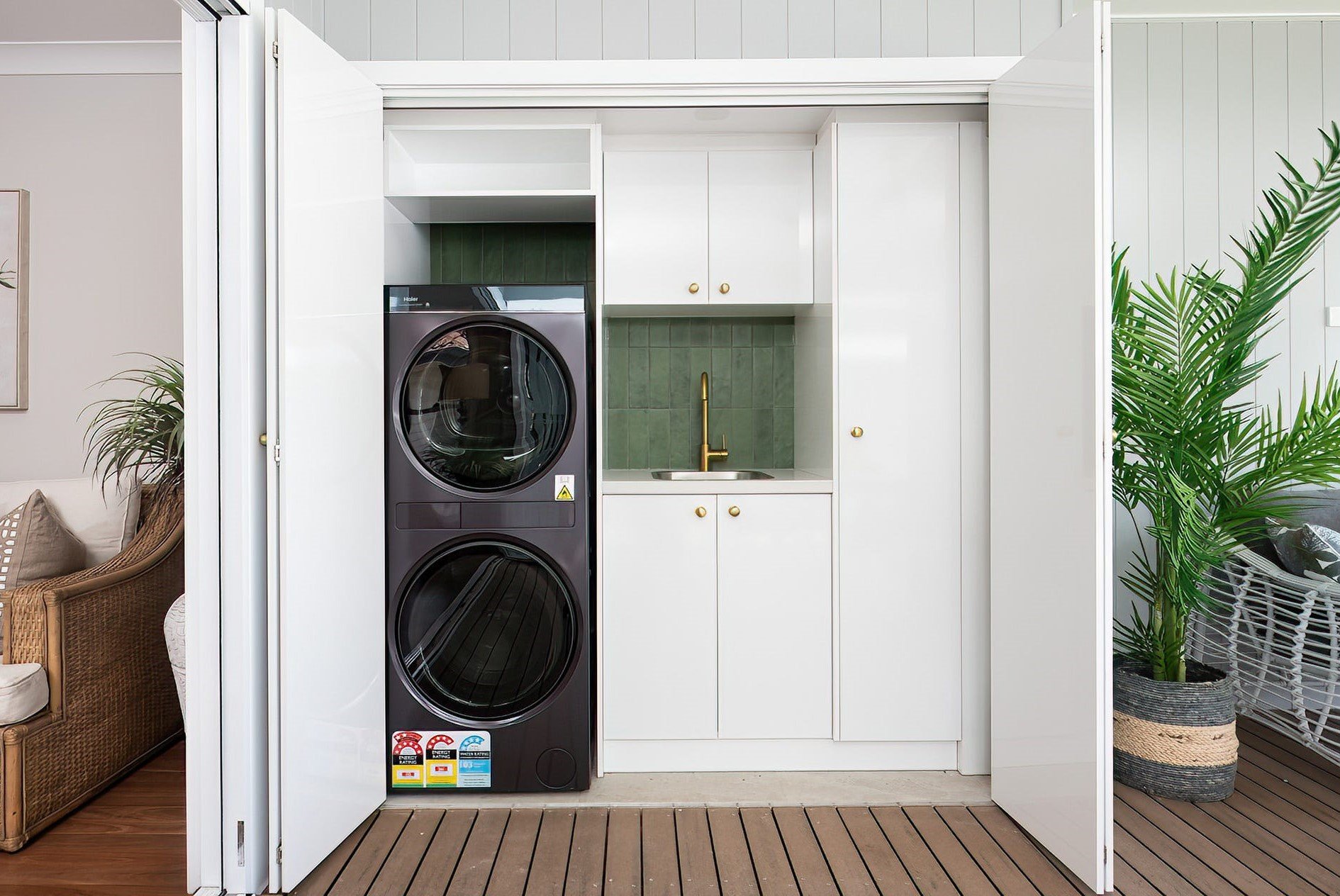 Laundry area with a stacked black washing machine and dryer, white cabinets, a small sink with a gold faucet, green tile backsplash, and outdoor wooden flooring, adjacent to a wicker sofa and large green plant.