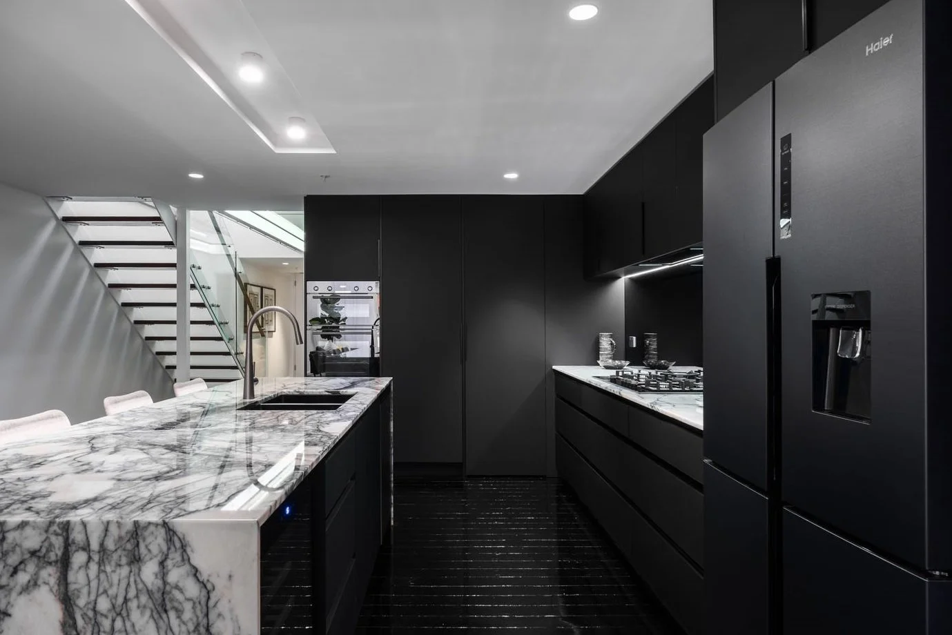 Modern black kitchen with marble countertops, black cabinets, and stainless steel appliances, including a large refrigerator with water dispenser, oven, and gas stove. There are recessed ceiling lights and a staircase in the background.