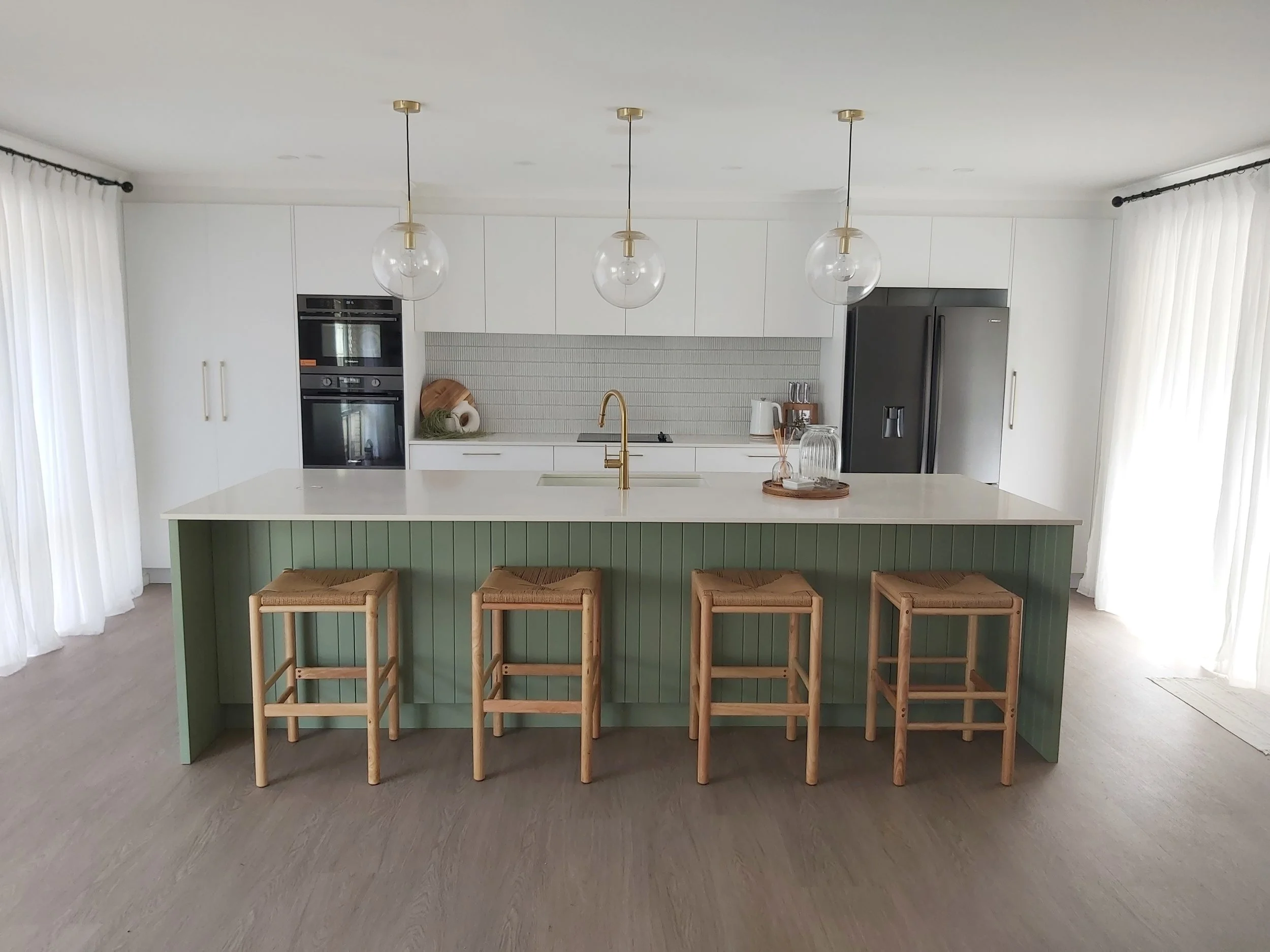 Modern kitchen with white cabinets, a large island with a green base, four wooden bar stools, pendant lights, a gray refrigerator, and a light-colored floor.