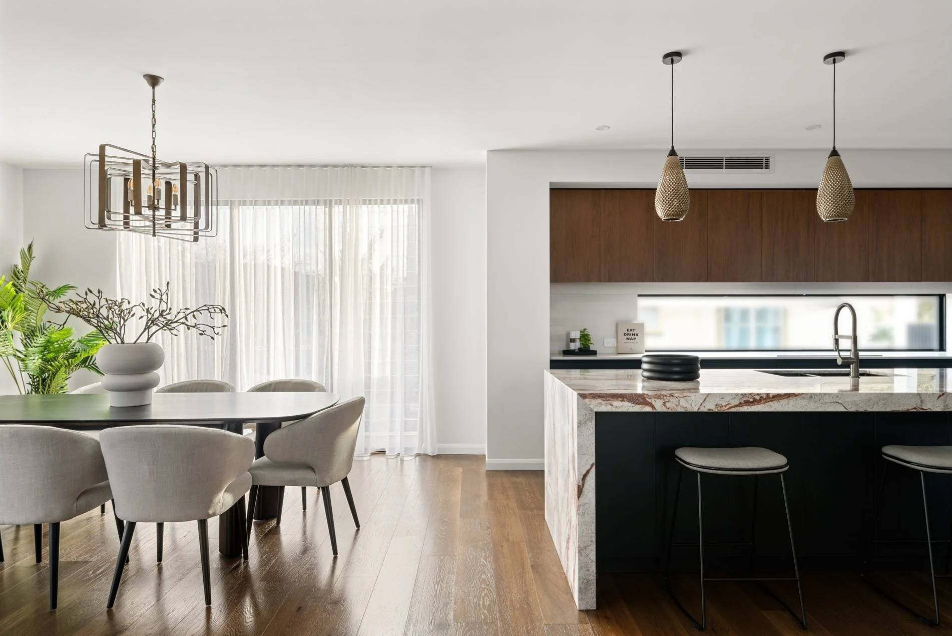 Modern open-concept kitchen and dining area with natural light, beige chairs around a dark dining table, a white vase with branches on the table, a marble kitchen island with two barstools, wooden cabinetry, pendant lights, and sheer curtains.