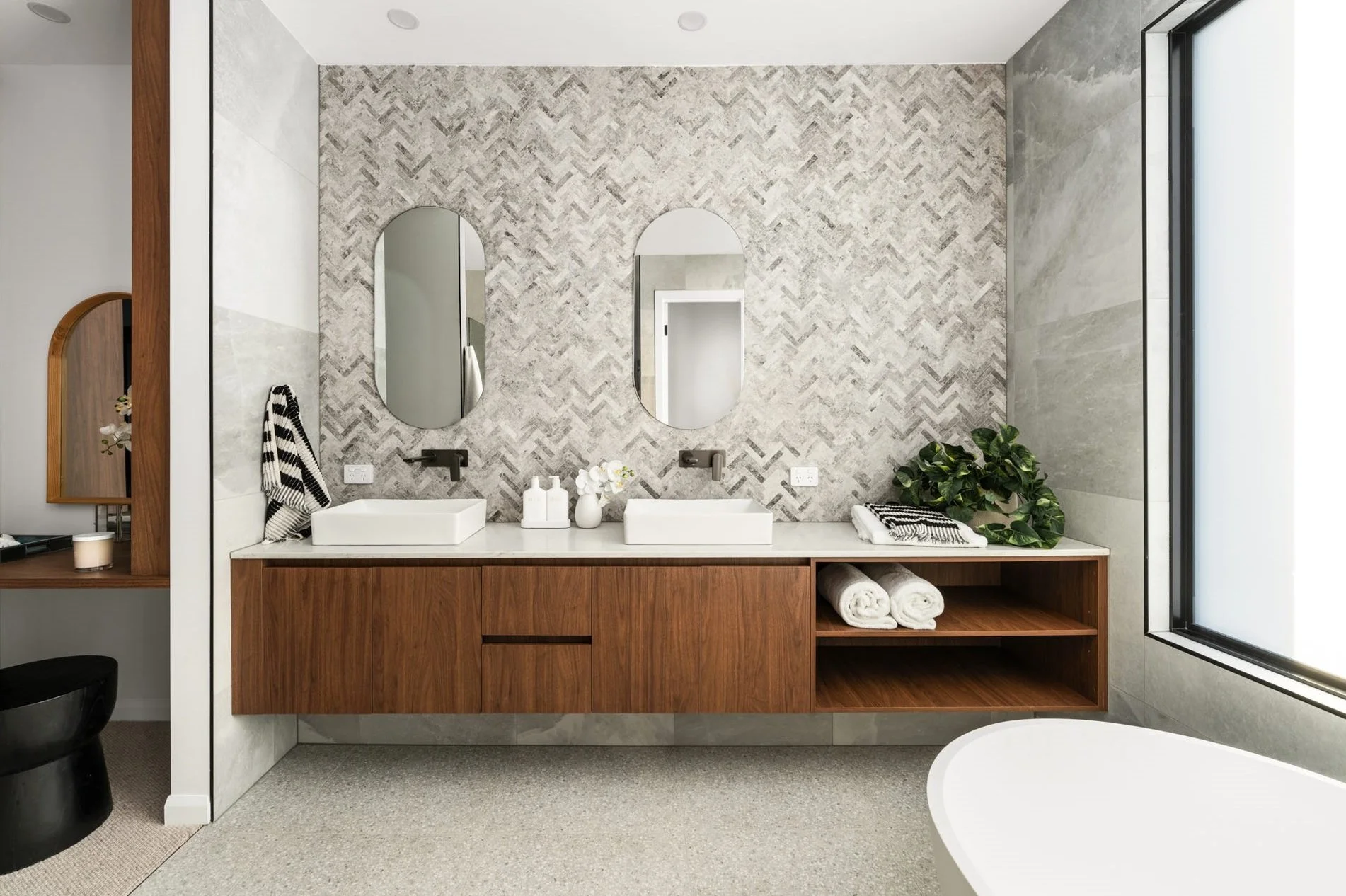 Modern bathroom with double sink, two oval mirrors, wooden vanity, white countertop, towels, decorative items, and large window.