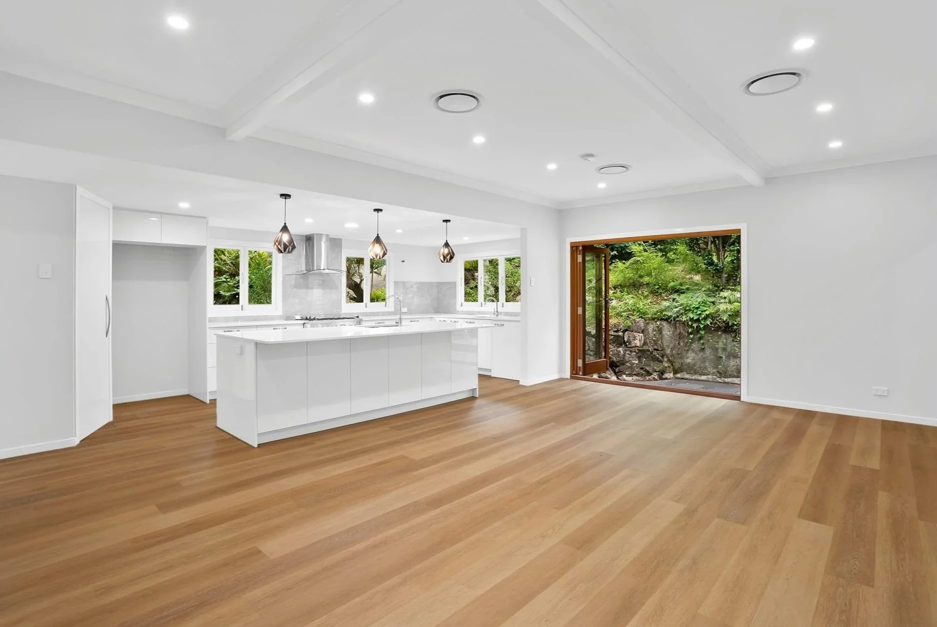 Bright, modern open-plan kitchen and living area with white walls, hardwood floors, large windows, and a sliding door opening to greenery outside.
