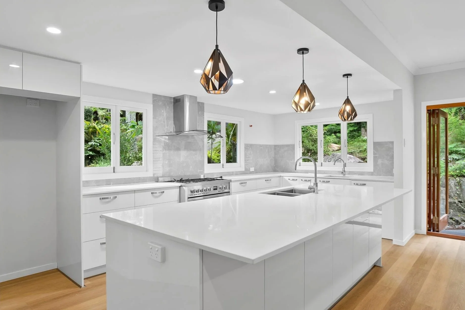 Modern white kitchen with large island, pendant lights, stainless steel appliances, and windows showing greenery outside.