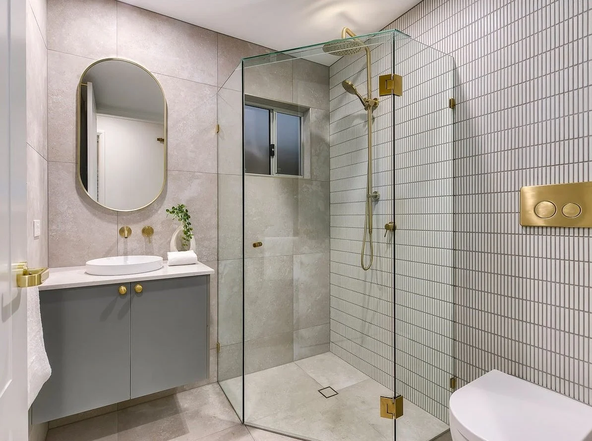 Modern bathroom with a walk-in shower enclosed by glass, brass fixtures, a small window, a gray vanity with a white vessel sink, an oval mirror, and a white toilet.