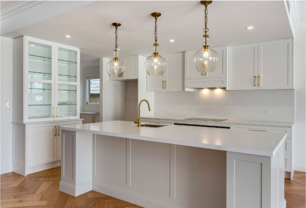 Modern kitchen with white cabinets, marble countertops, and a large island with a gold faucet. Three pendant lights hang above the island, and there is a glass-front cabinet to the left. Hardwood floors and additional cabinetry are visible.