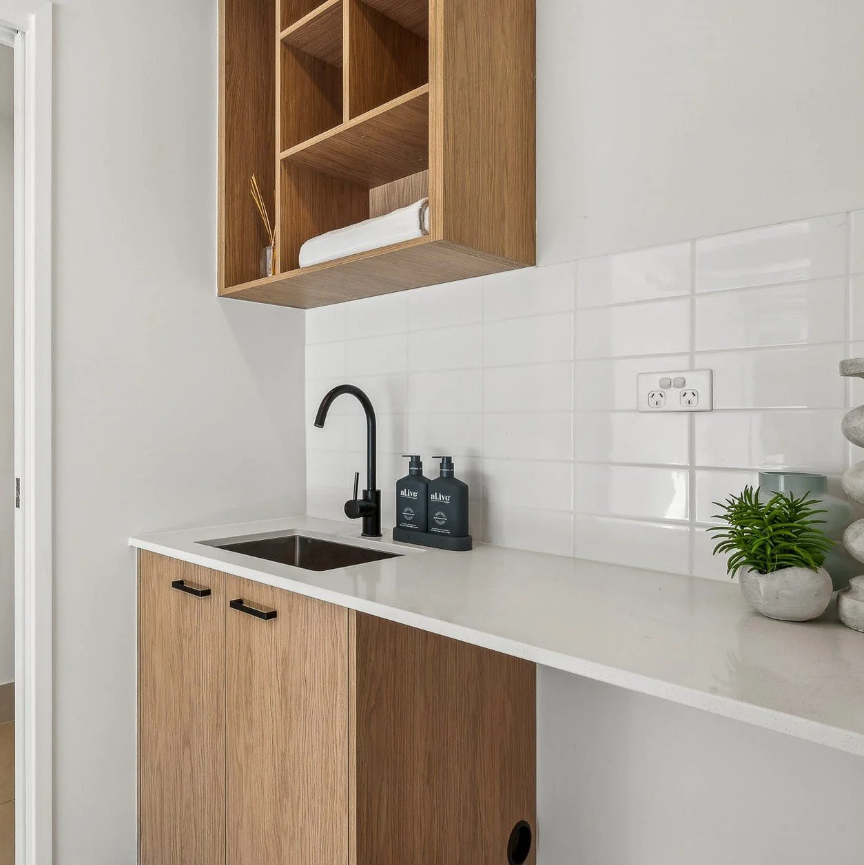 Kitchen area with a white countertop, black faucet, two black soap dispensers, wooden cabinets, white tile backsplash, electric outlets, a small green potted plant, and a stack of smooth stones.