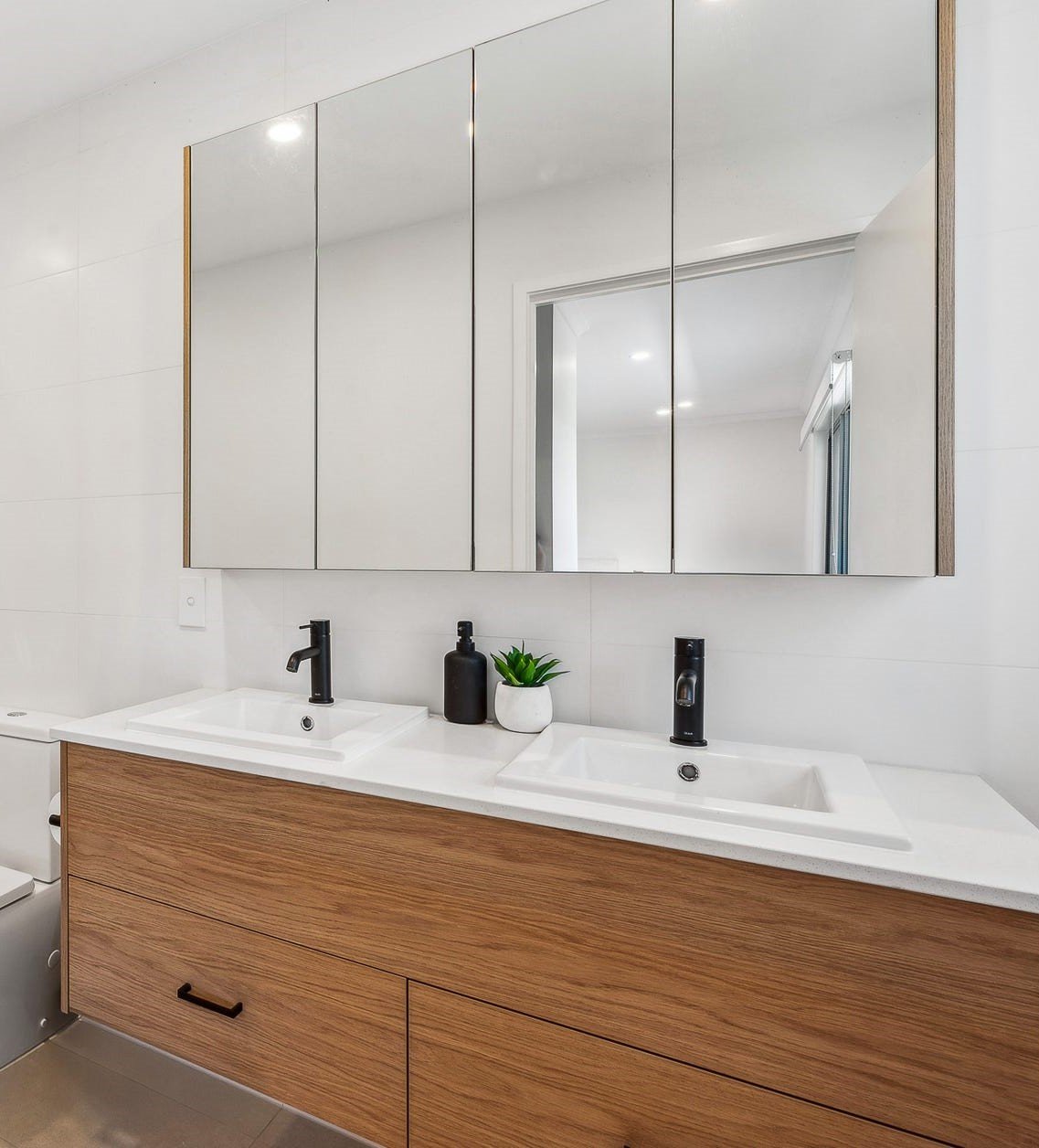 Modern bathroom with double vanity, white countertop, black faucets, mirrored cabinet, and decorative plants.
