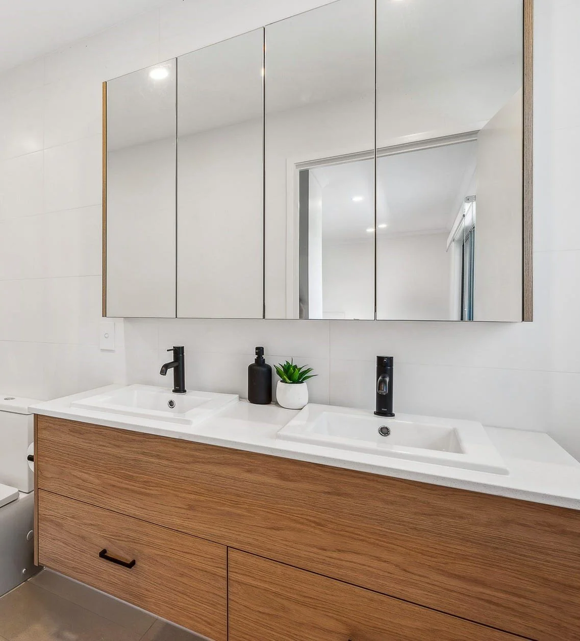 Modern bathroom vanity with dual sinks, black faucets, a large mirror, a small green plant in a white pot, and a black soap dispenser, with a wooden cabinet and neutral-colored walls.