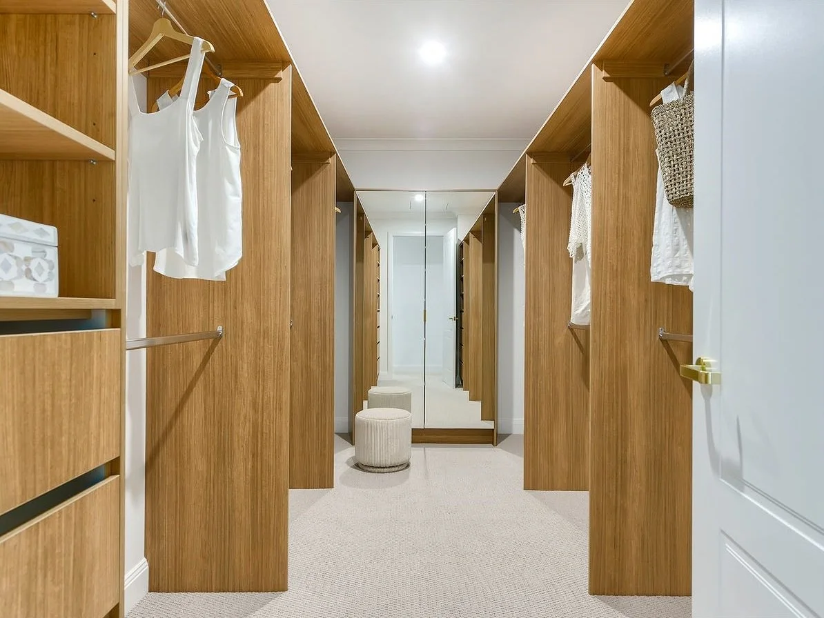 Walk-in closet with wooden shelving and hanging clothes, with mirror at the back, a small beige stool, and a white door on the right