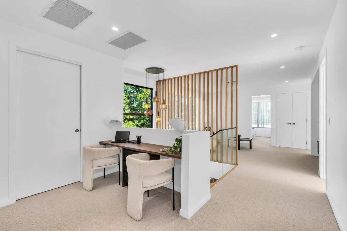 Bright, modern living space with a small workspace, beige chairs, wooden slat divider, and greenery outside window.