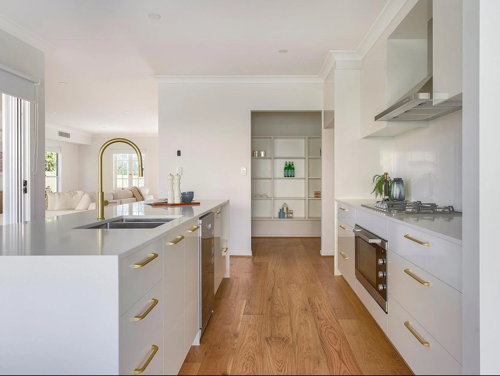 Modern kitchen with white cabinets, gold handles, wooden flooring, and built-in appliances, leading to a living area with a sofa and windows.
