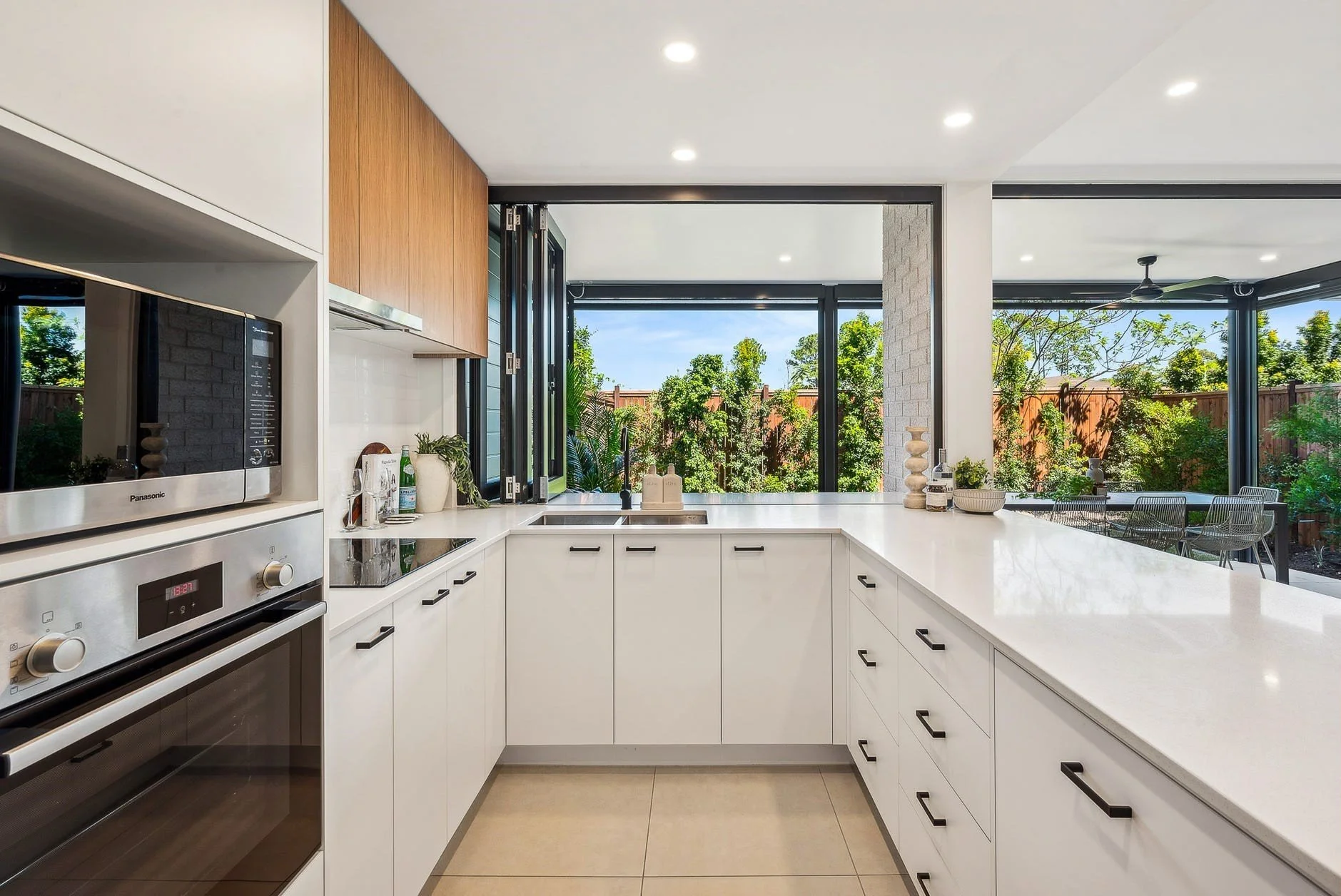 Modern kitchen with white cabinets, black hardware, and a view of a lush backyard through large open windows.