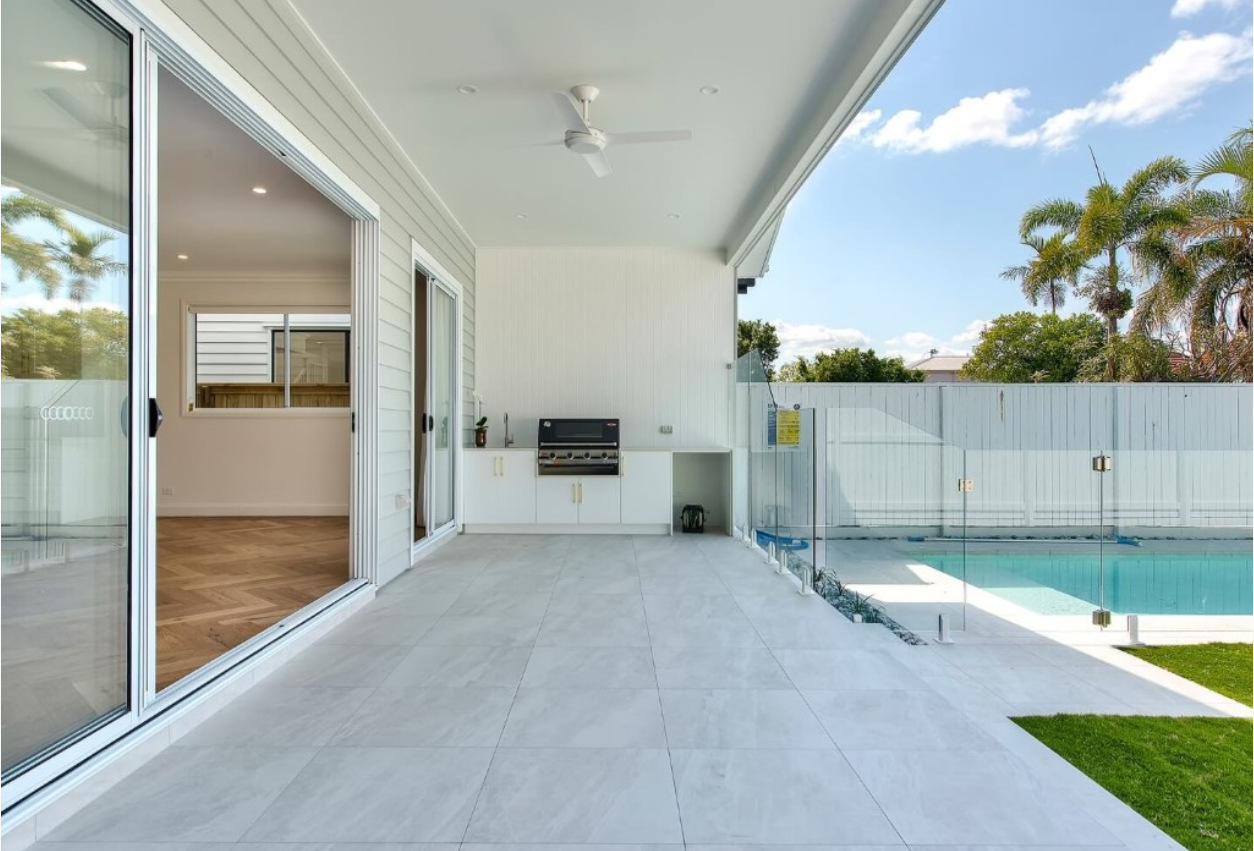 Modern backyard patio with sliding glass doors, ceiling fan, and swimming pool enclosed by glass fence, surrounded by tropical trees and blue sky.