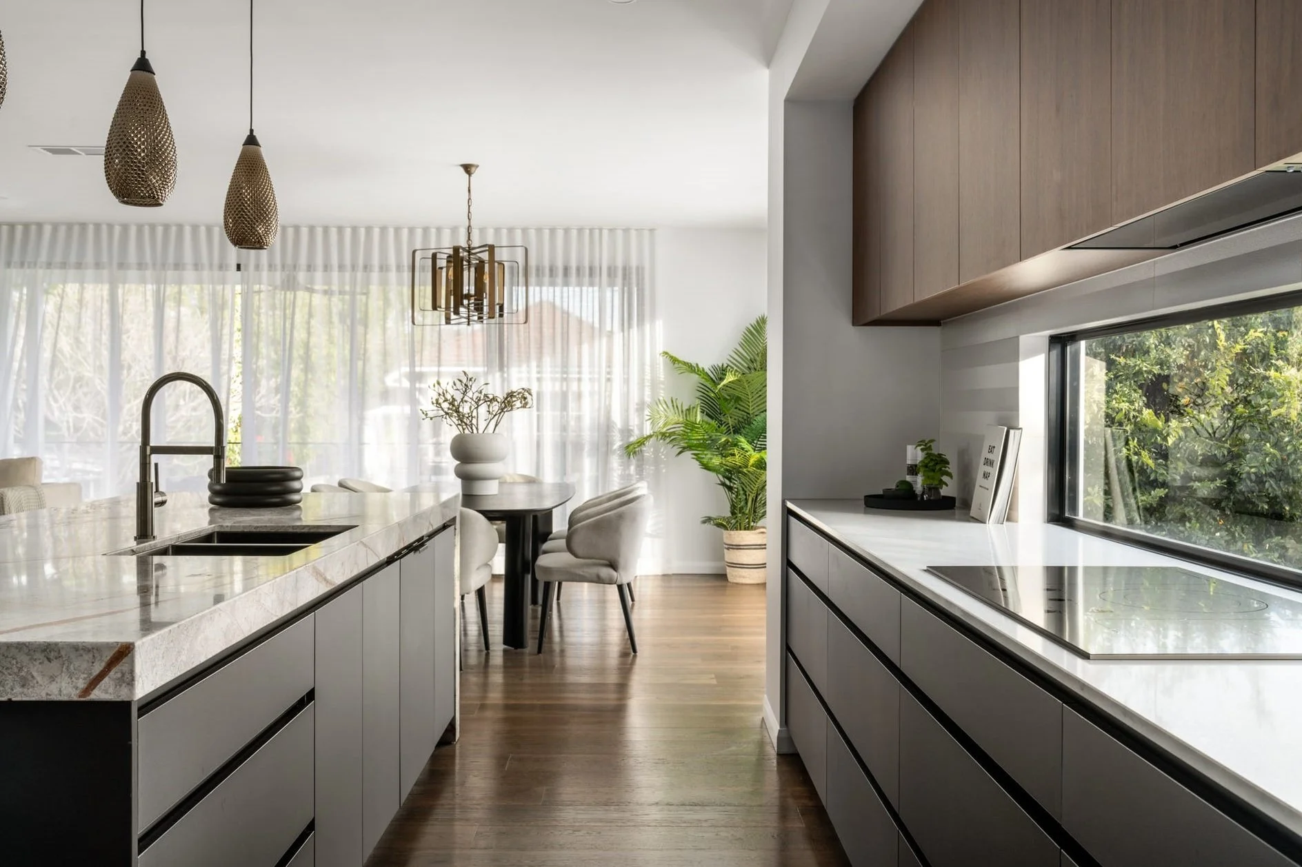 Modern kitchen with marble countertop island, pendant lights, and a dining area with chairs and a potted plant near large windows with sheer curtains.