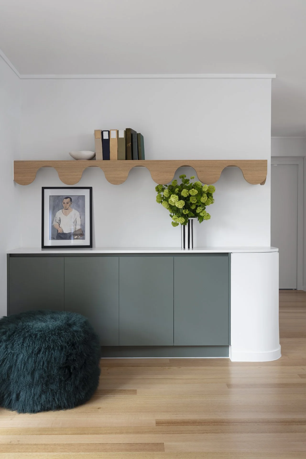 Interior of a home with a green cabinet, a fluffy dark green pouf, a framed portrait, a vase of green flowers, a wooden shelf with books, and a white wall.