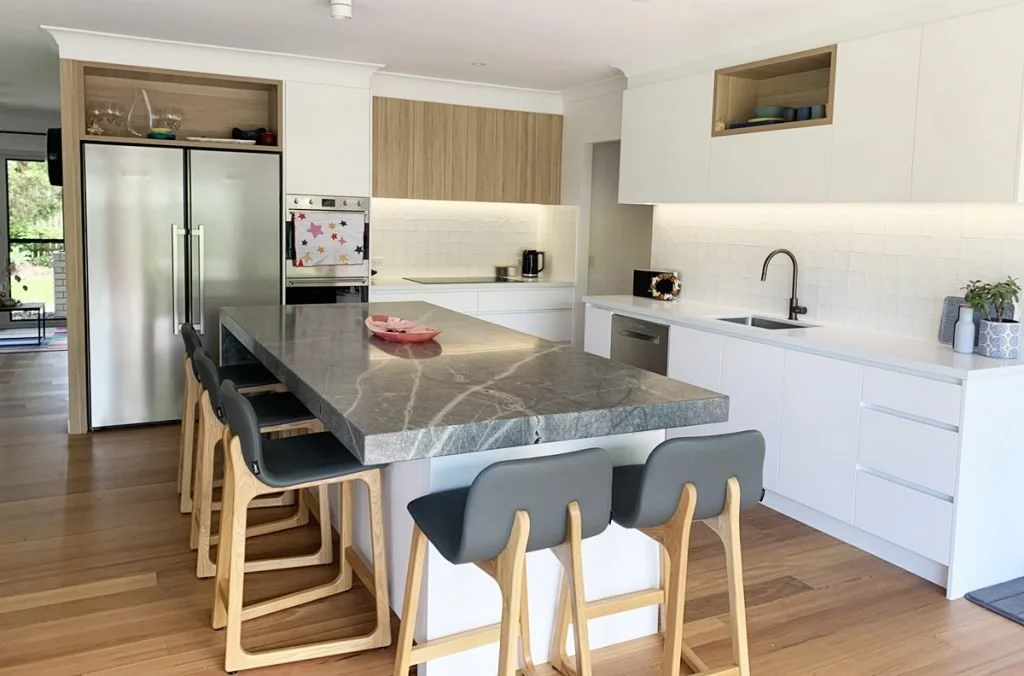 Modern kitchen with white cabinetry, a large marble island with black and brown veining, four black and gray bar stools with wooden legs, stainless steel refrigerator, built-in oven with colorful star magnets, open shelving with dishes, and a small p