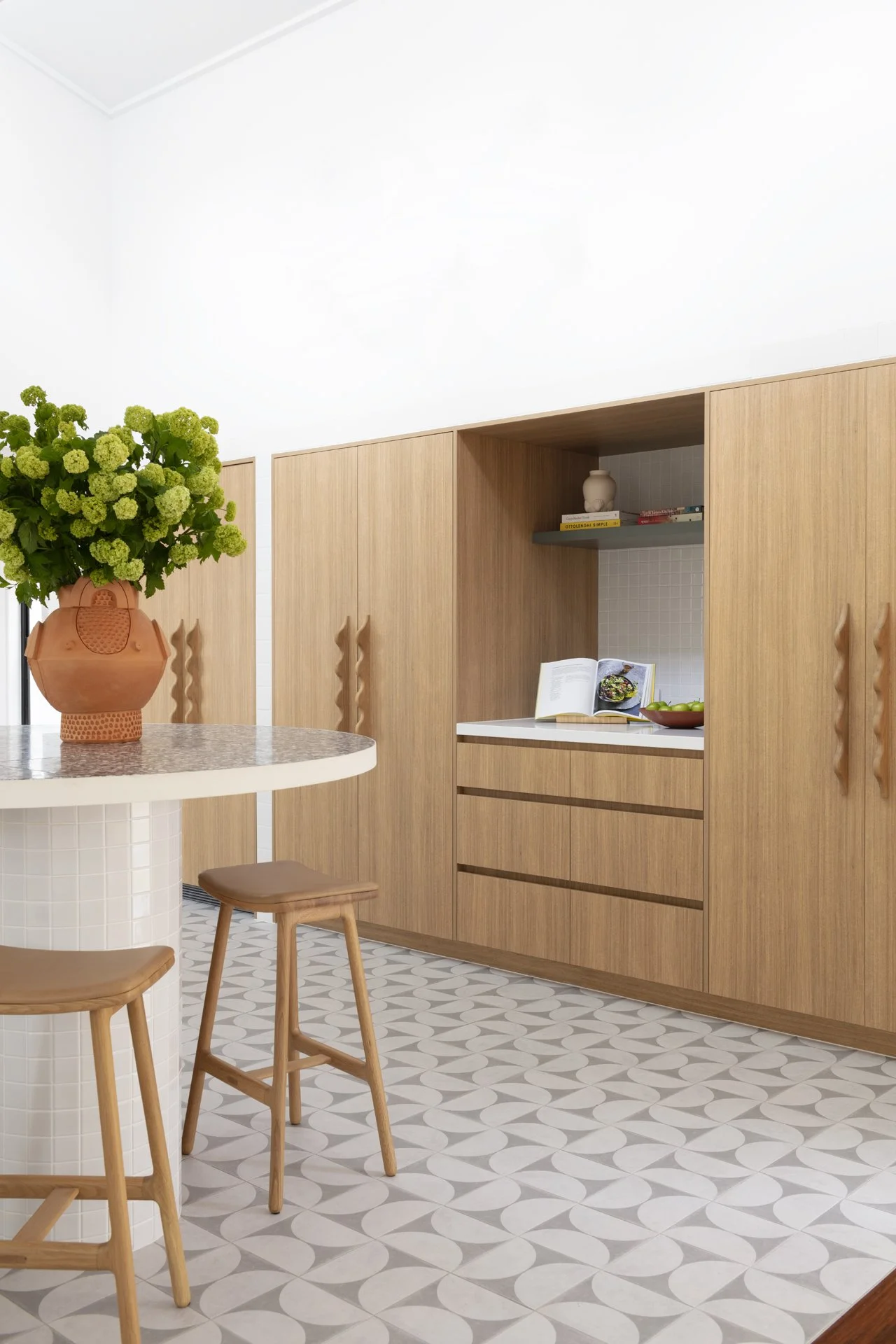 A modern kitchen with white walls, light wood cabinets, a white countertop, and patterned tile flooring. A vase with green hydrangeas is on a round marble-top table with wooden stools, and an open cookbook and a bowl of green apples are on the counte