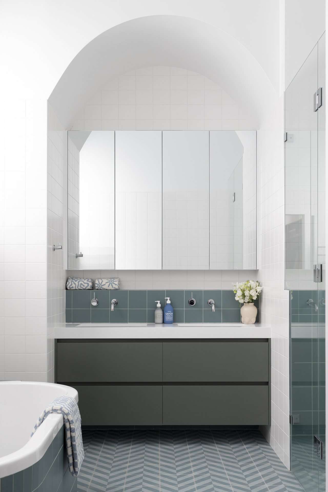 Modern bathroom with a large mirror, white tiled walls, a gray vanity with two drawers, a white sink, and a green tiled backsplash. Bathroom accessories include soap dispensers, rolled towels, a vase with white flowers, and a towel hanging on the sid