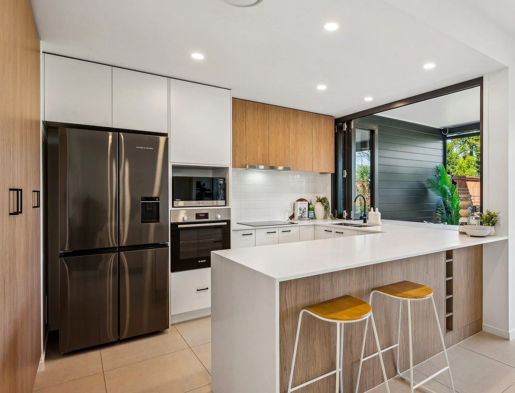 Modern kitchen with stainless steel refrigerator, built-in microwave and oven, white and wood cabinets, open window looking to outdoor patio with plants