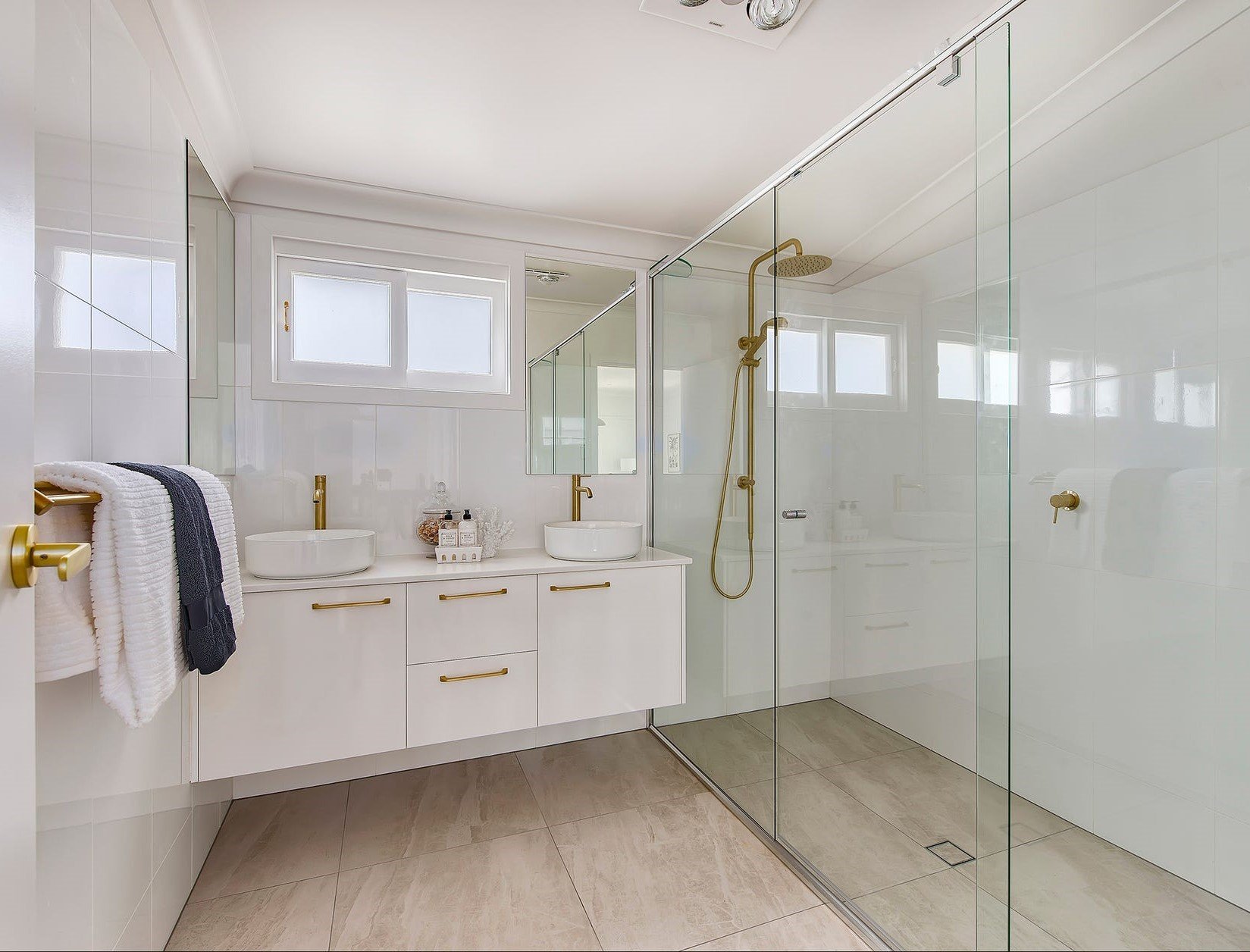 Modern bathroom with white cabinetry, double vessel sinks with gold fixtures, and a walk-in glass shower with gold shower head. Two small frosted windows let in natural light, and there are beige tiled floors.