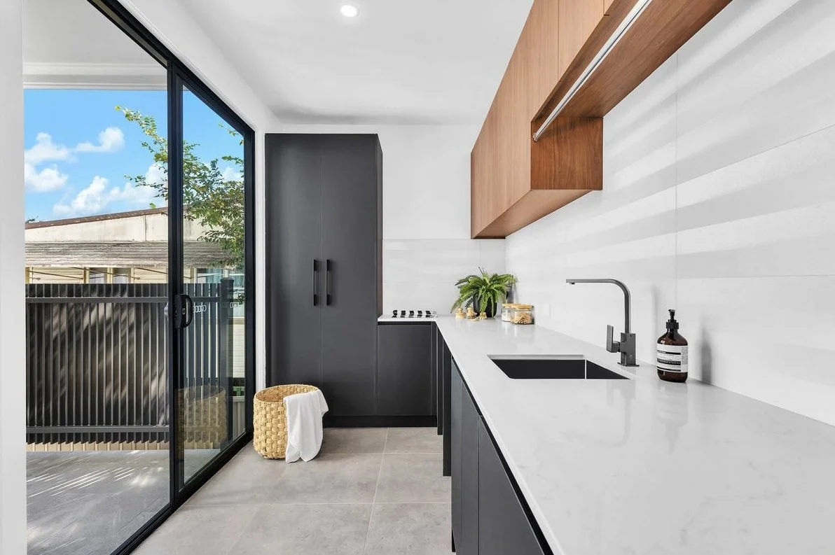 Modern kitchen with black lower cabinets, a white countertop, a black sink, and wooden upper cabinets. It has a large sliding glass door leading to a balcony with a black railing and a view of a tree and blue sky.
