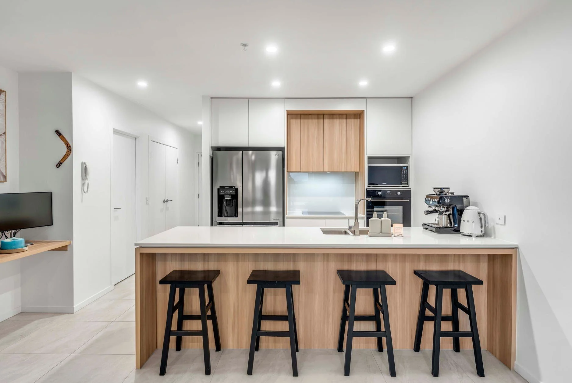 Modern kitchen with white walls, wooden accents, a stainless steel refrigerator, microwave, oven, coffee machine, and four black bar stools at a white countertop island.