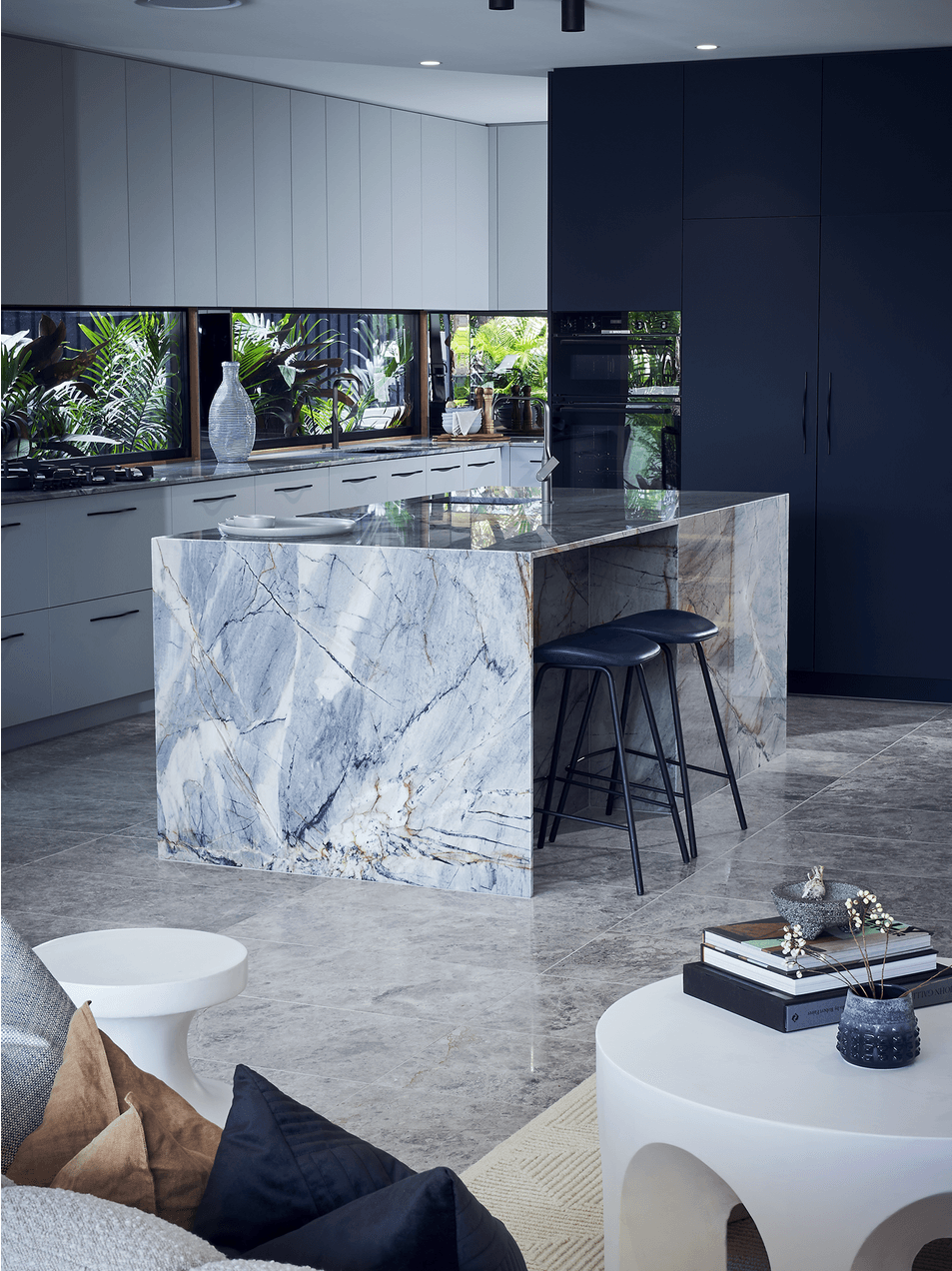 Modern kitchen with a marble island, white and dark blue cabinets, and large windows with tropical plants outside.
