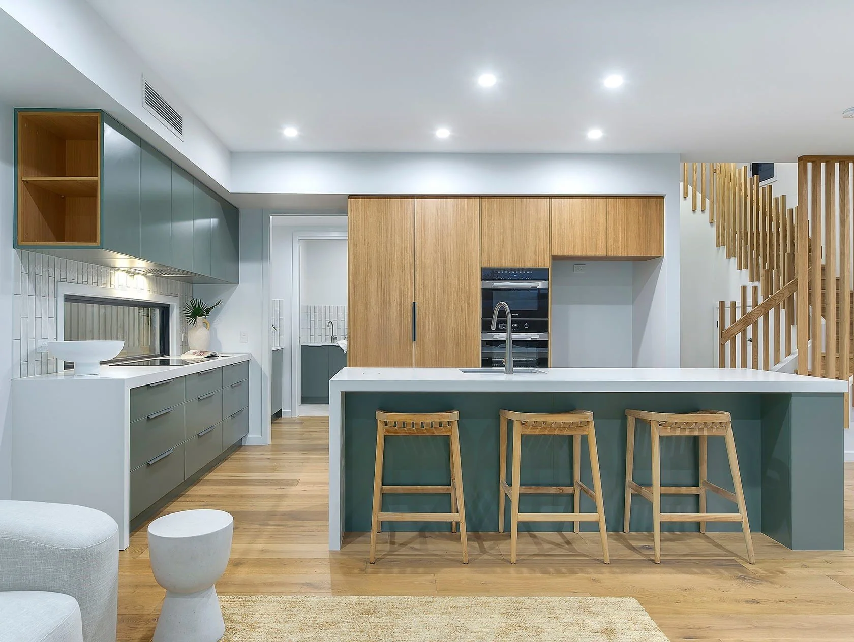 Modern kitchen with wooden and green cabinets, white countertops, three wooden bar stools, a sink, and a staircase with wooden railing.