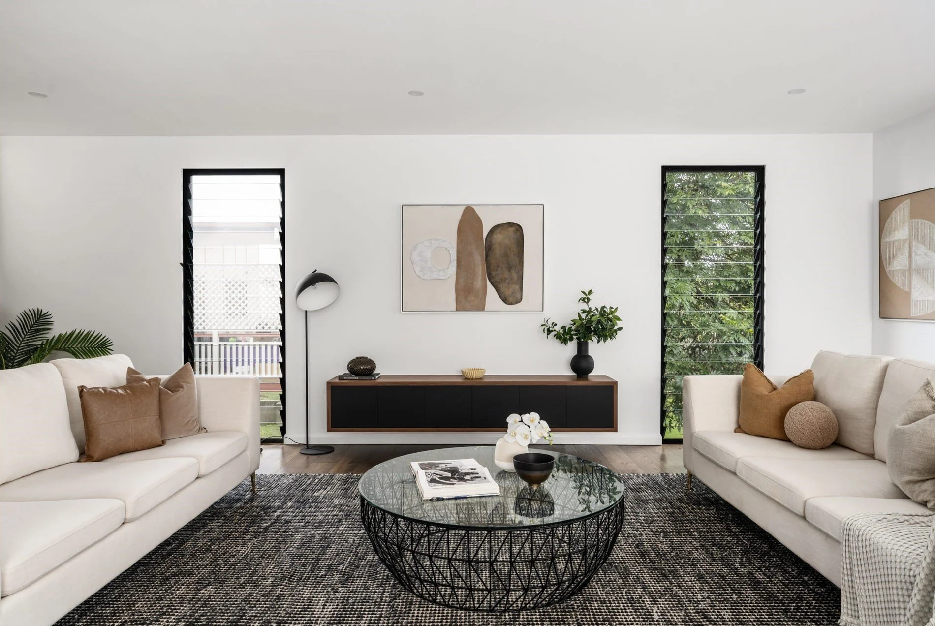 Modern living room with white sofas, a black coffee table with a glass top, a wall-mounted console, abstract wall art, and large windows with black louvered blinds.
