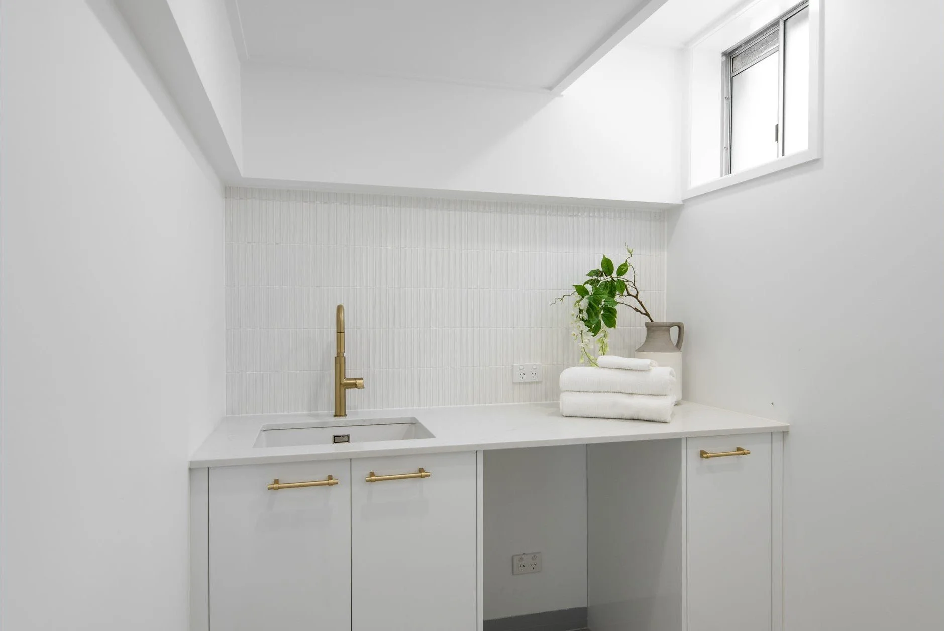 Minimalist white laundry or utility room with a countertop, gold handles, a brass faucet, stacked towels, a potted plant, and a small window.