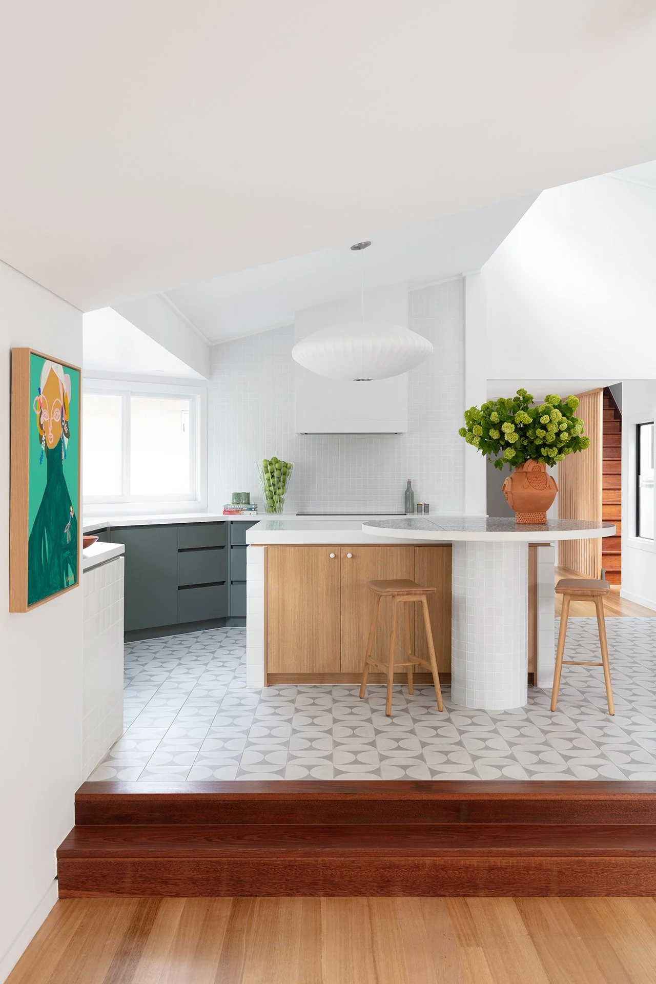Bright modern kitchen with wood accents, white tiled backsplash, gray and wood cabinetry, patterned tile flooring, and a large green floral arrangement on the island.