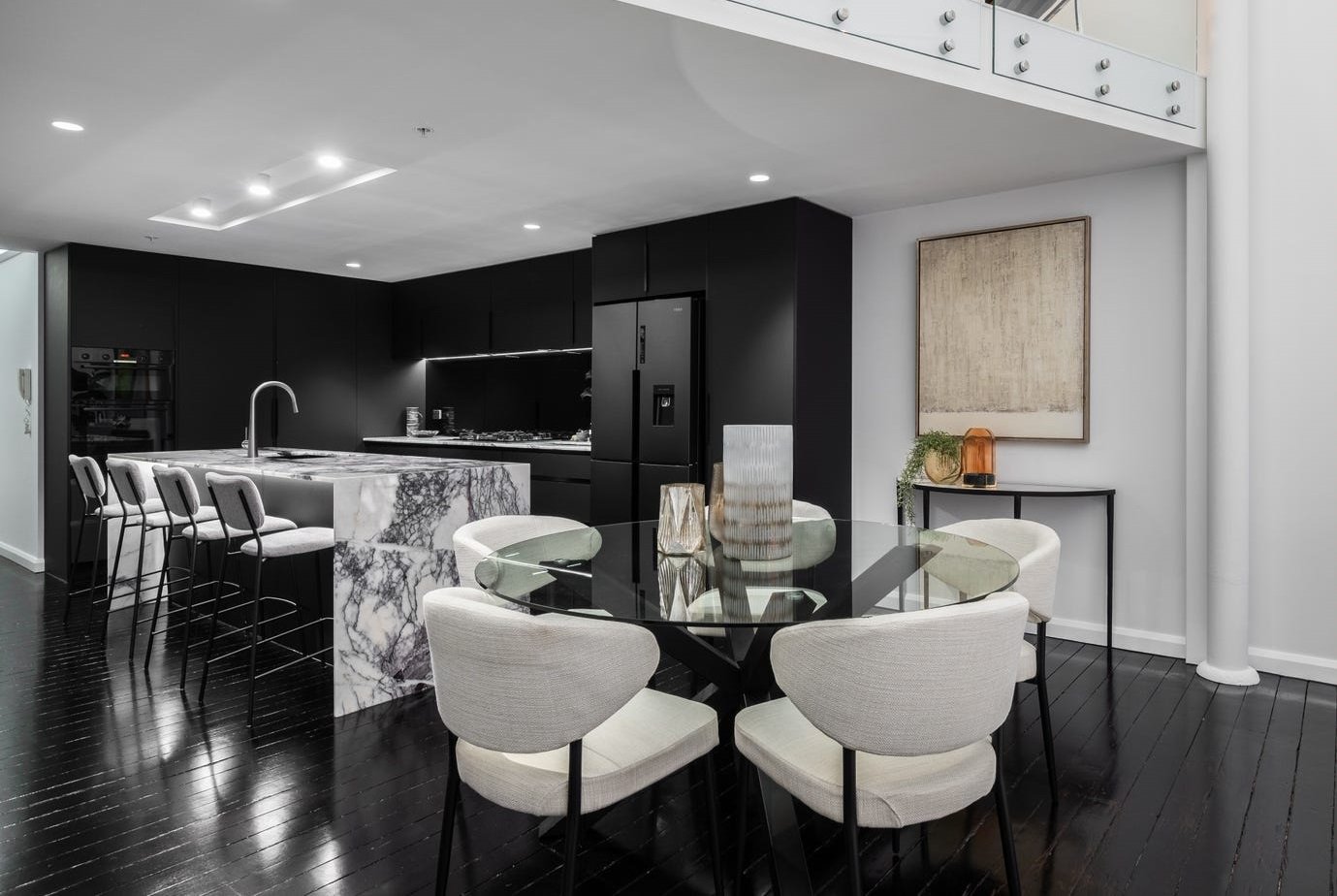 Modern kitchen and dining area with black cabinetry, marble island, glass table, and beige chairs on dark hardwood floors.