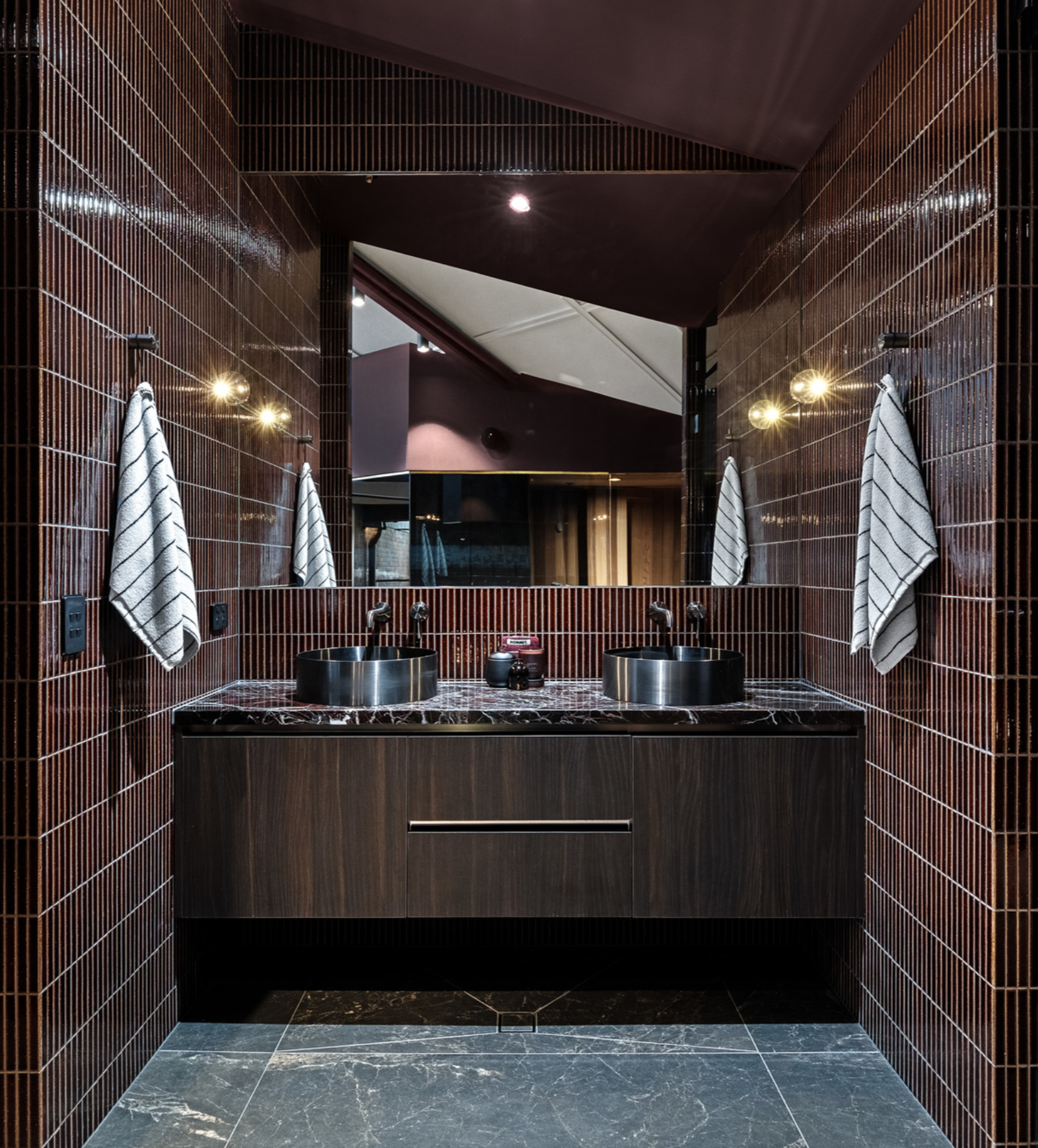 Modern bathroom with dark red tiled walls, a large mirror, two black vessel sinks, and black and white striped towels hanging on either side.
