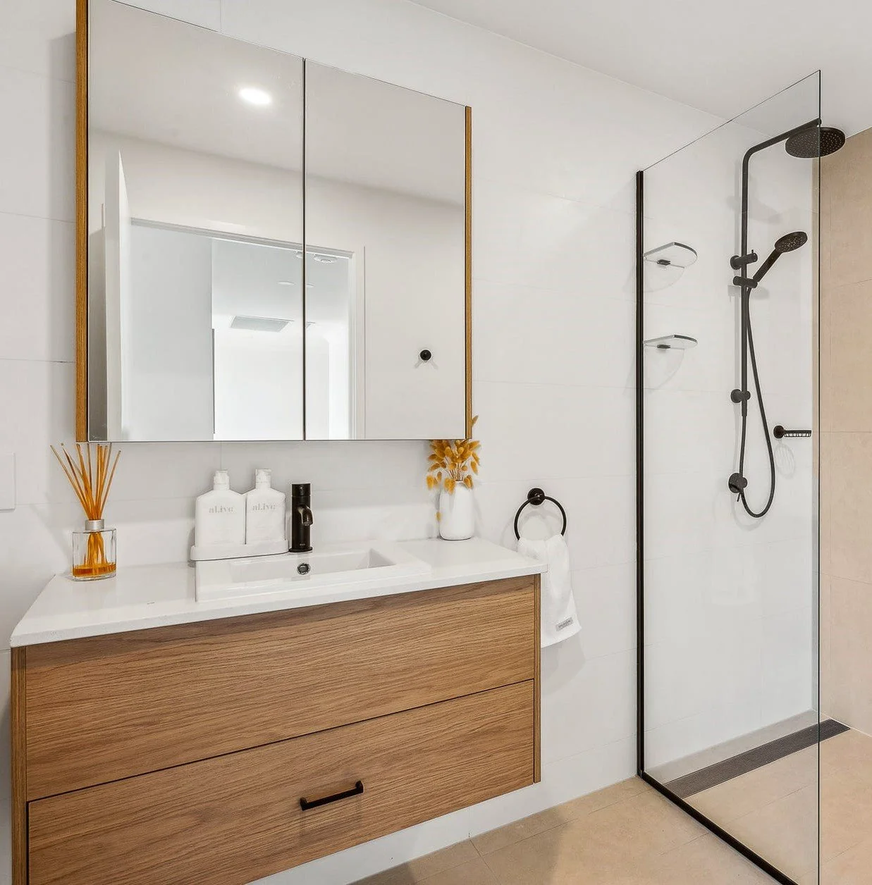 Modern bathroom with a wooden vanity, white countertop, and a large mirror. There is a glass shower on the right with black fixtures and a beige tile wall.