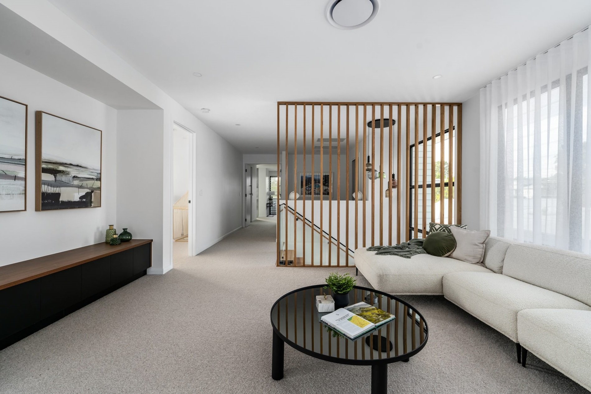 Living room with a white sectional sofa, black coffee table with magazines, light-colored carpet, and natural light through large windows with sheer curtains. Decorative wall art and a wooden slat partition near the staircase are visible.