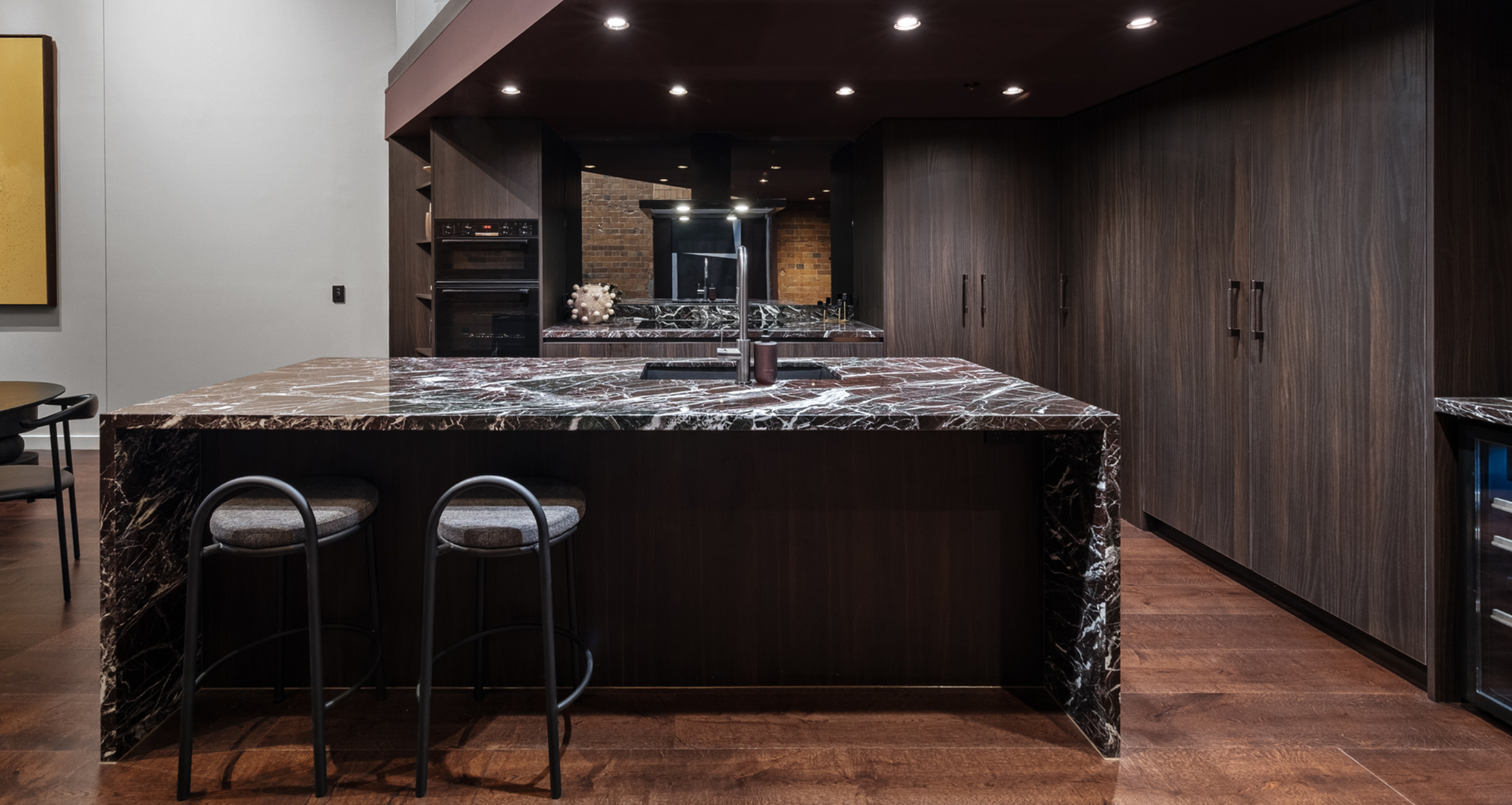 Modern kitchen with dark wooden cabinets, a marble countertop island with a sink, and wooden flooring. There are two barstools at the island, and recessed ceiling lights.