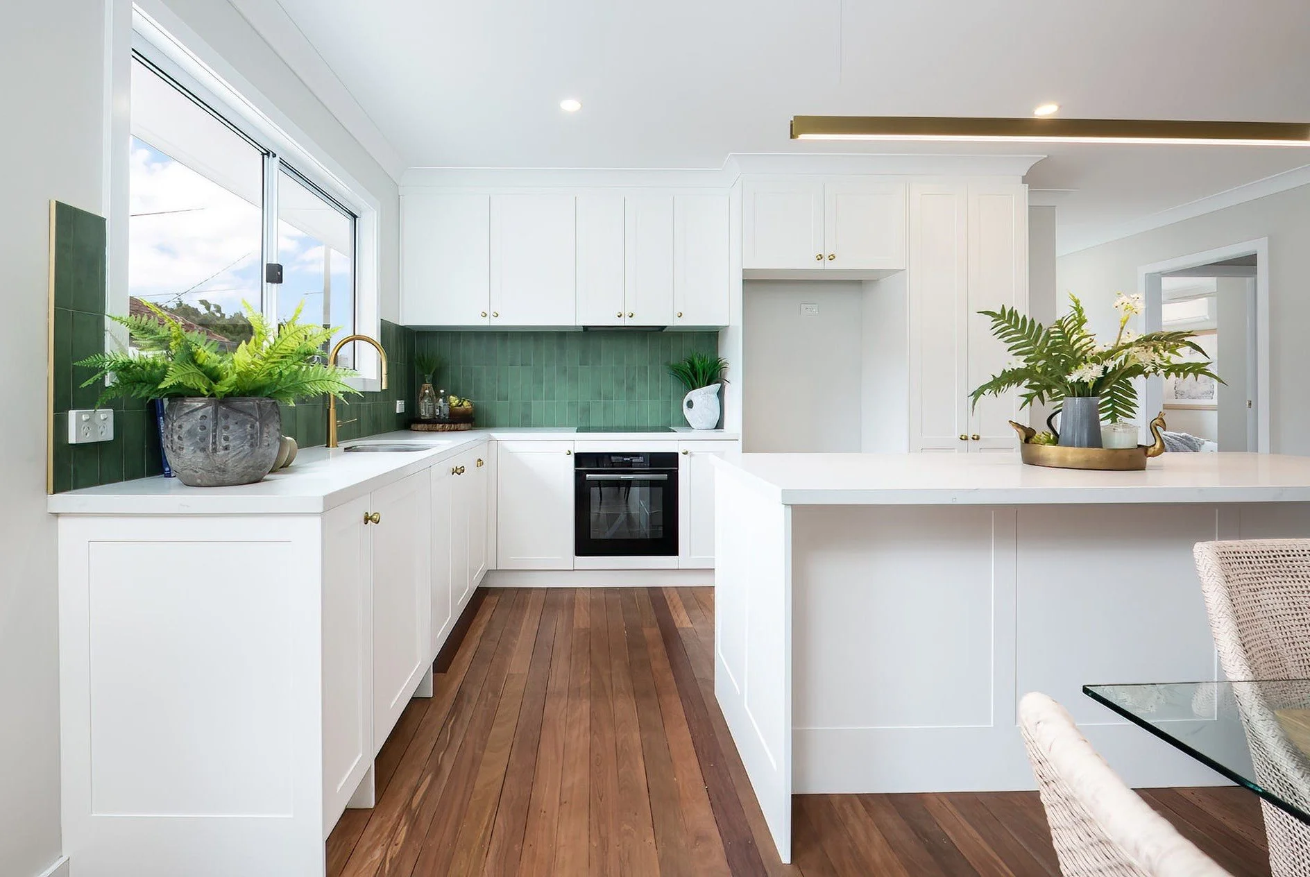 Modern kitchen with white cabinetry, green tile backsplash, wooden floor, large window, and decorative plants.