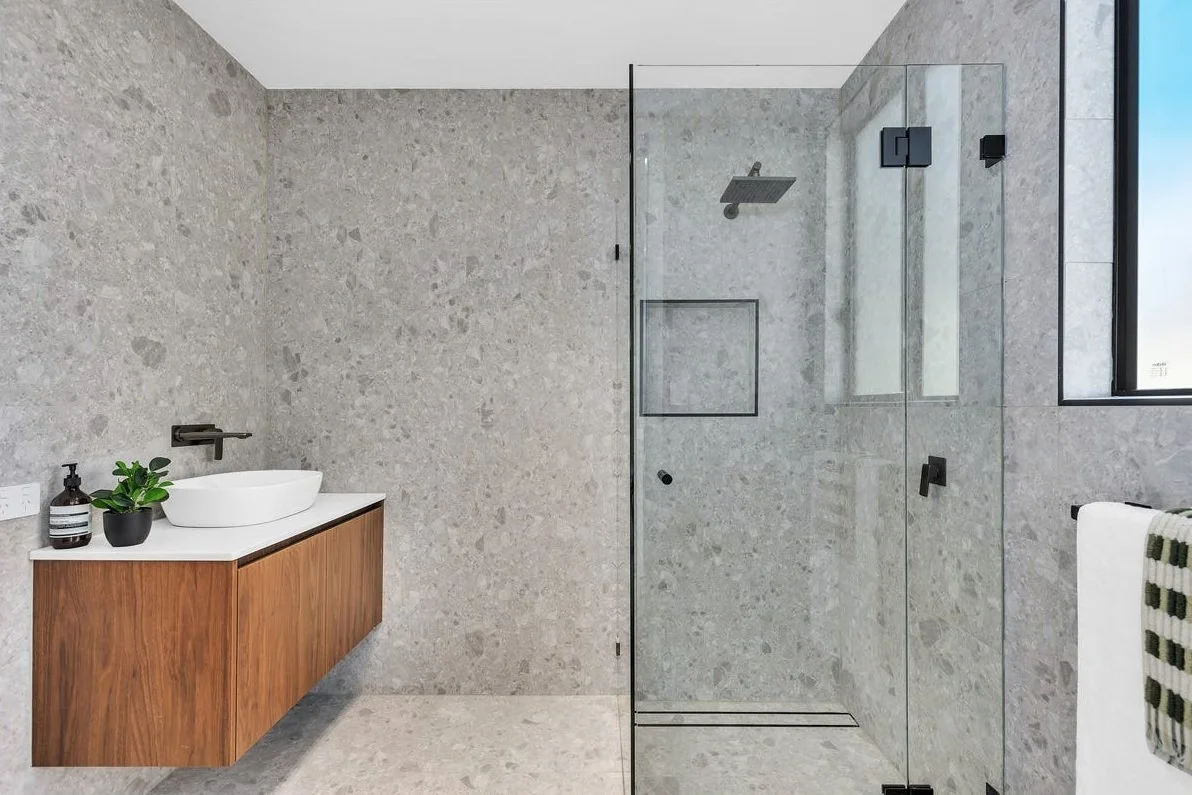 Modern bathroom with a glass-enclosed shower, gray stone walls, and a floating wooden vanity with a white vessel sink and black faucet, along with a plant and soap dispenser on the counter.