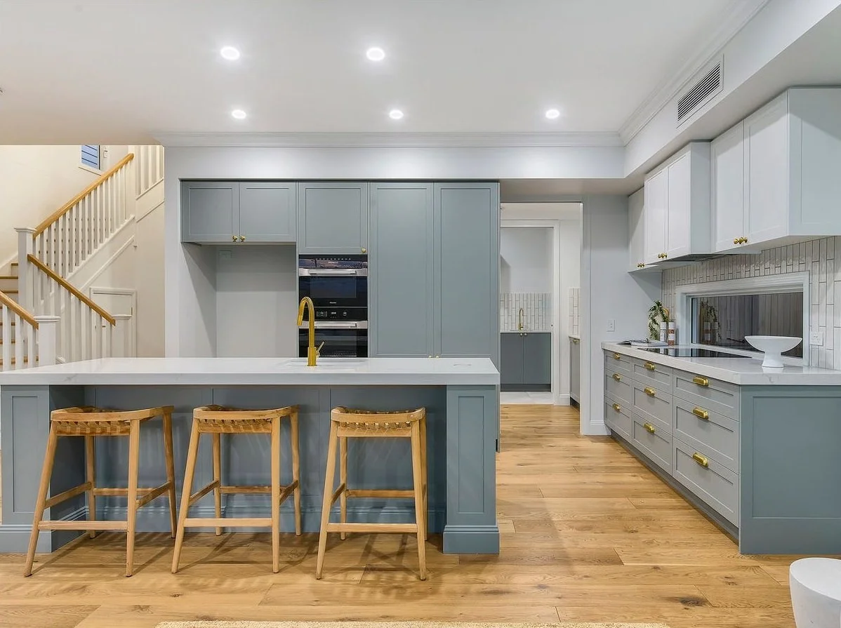 Modern kitchen with light blue and white cabinets, wooden countertops, three wooden barstools, and hardwood flooring.