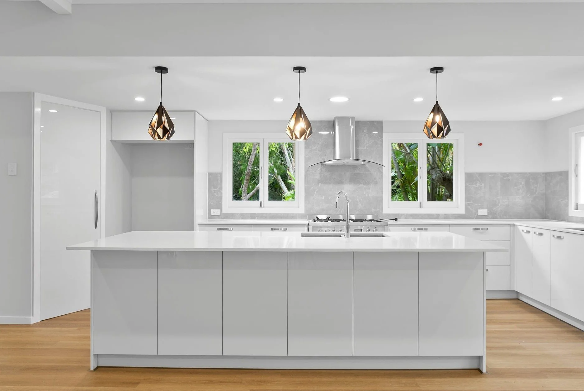 Modern white kitchen with island, pendant lighting, gray marble backsplash, and green trees visible through windows.
