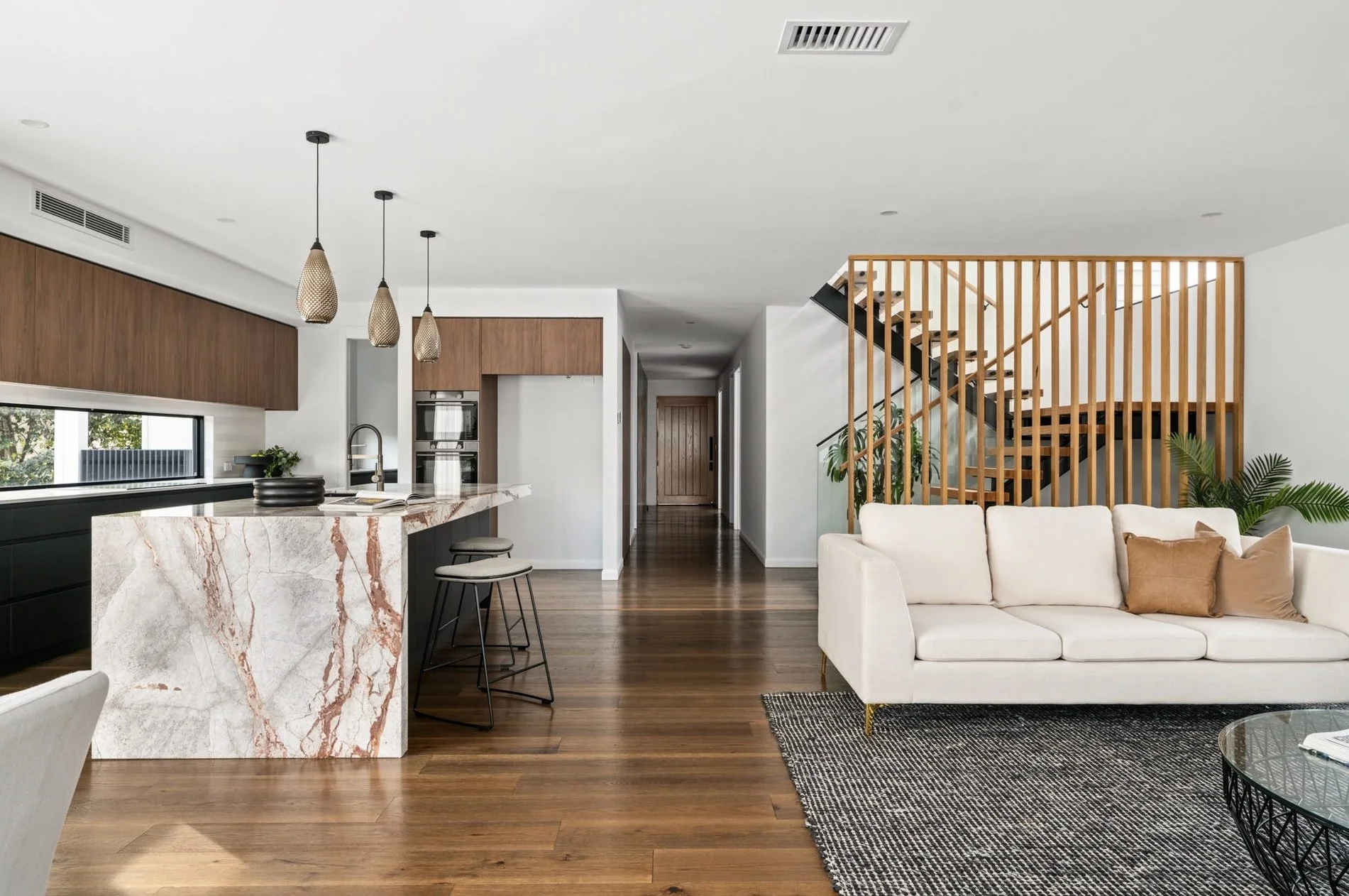 Modern open-concept living room and kitchen with wooden flooring, white walls, a staircase with wooden slats, a white sofa with brown and beige pillows, a black and white rug, plants, and a marble kitchen island with bar stools.