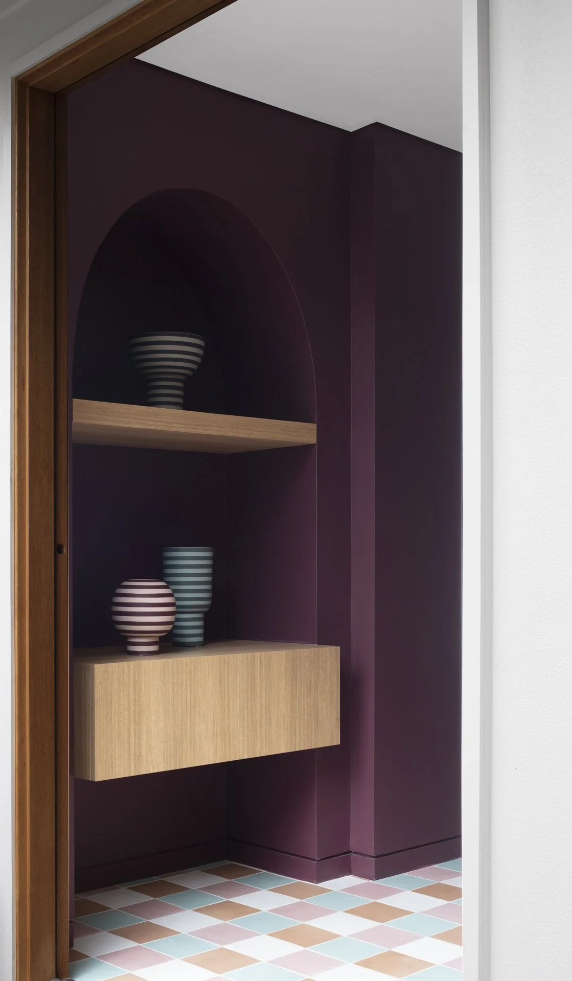 A built-in wall niche with a dark purple interior and wooden shelves containing striped vases, with a partial view of a doorway and colorful tiled floor.