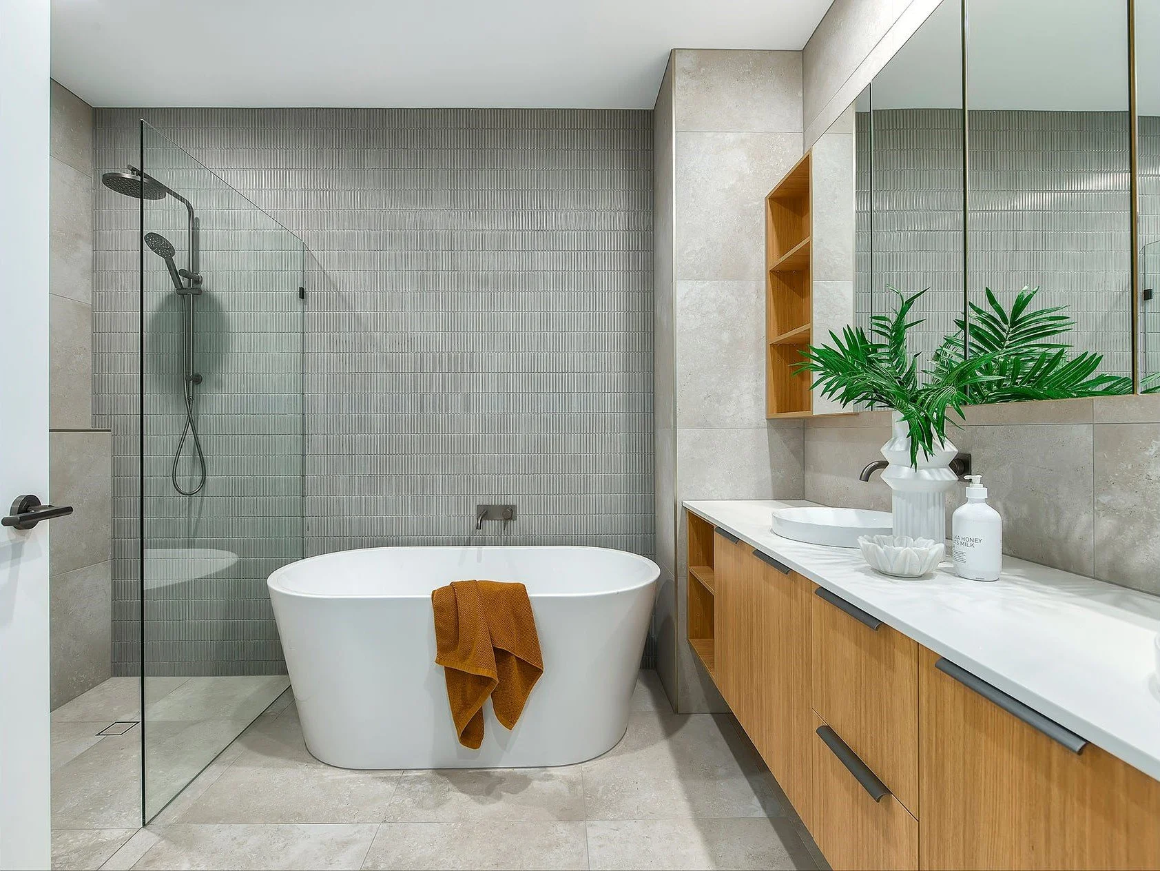 Modern bathroom with a walk-in shower, freestanding bathtub, wooden vanity with a white countertop, large mirror, plant, and decorative items.