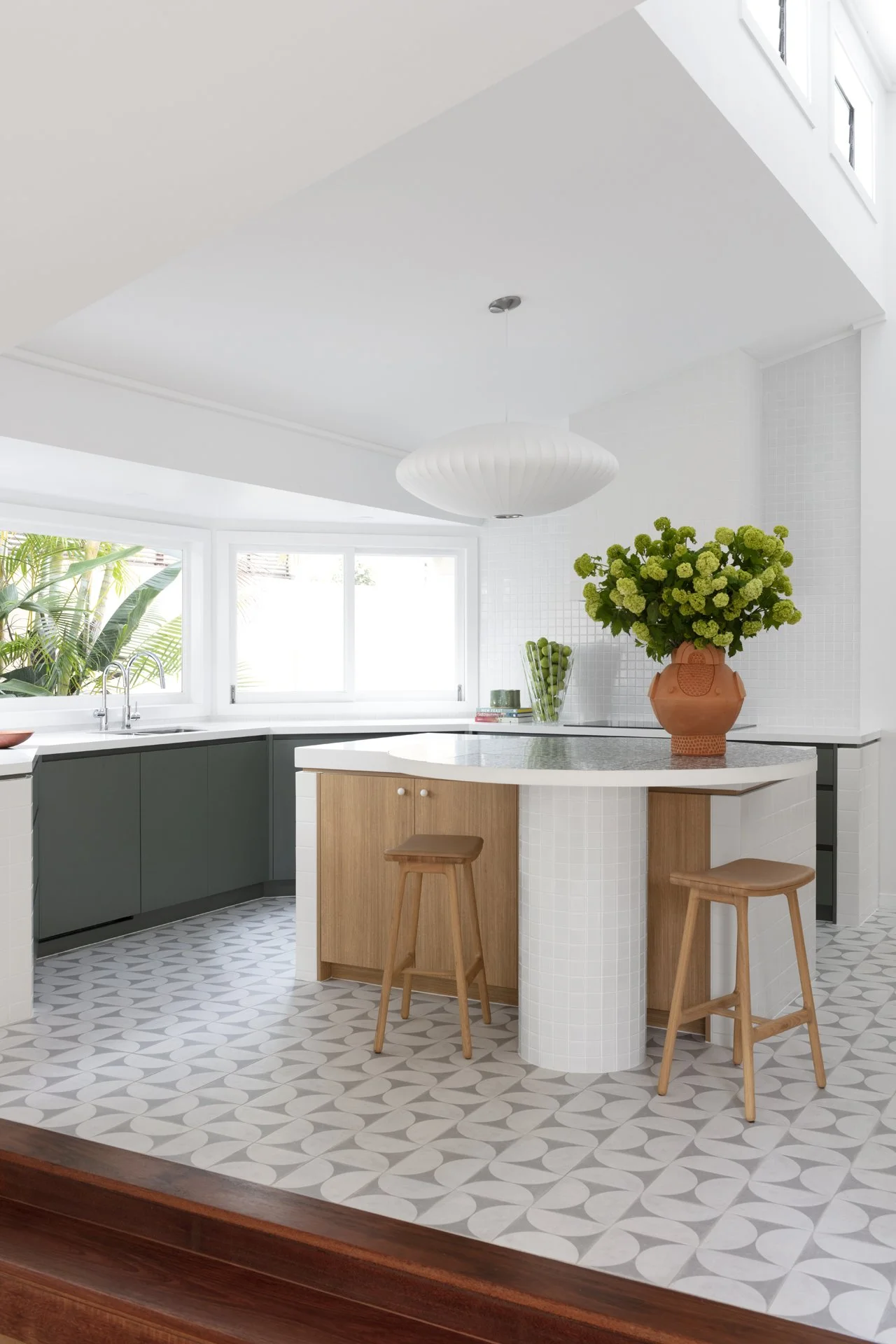 Modern kitchen with green lower cabinets, white upper walls, a central island with wooden panels, a large terracotta vase with green flowers, patterned floor tiles, and large windows letting in natural light.